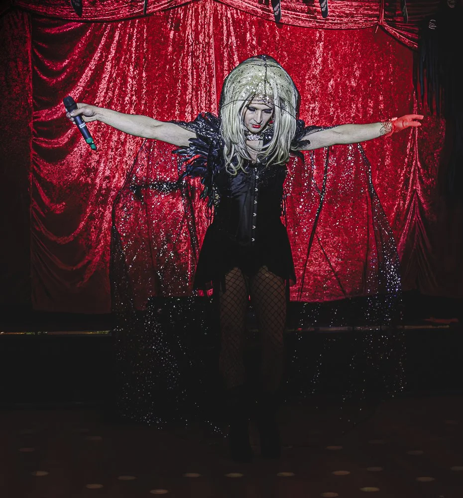 Performer on stage with arms outstretched, wearing a black dress, fishnet stockings, and a light-colored wig, in front of red velvet curtains, holding a microphone.
