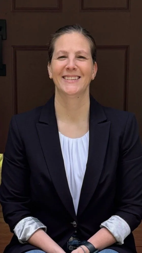A woman smiling, wearing a black blazer over a white blouse, seated in front of a brown door.