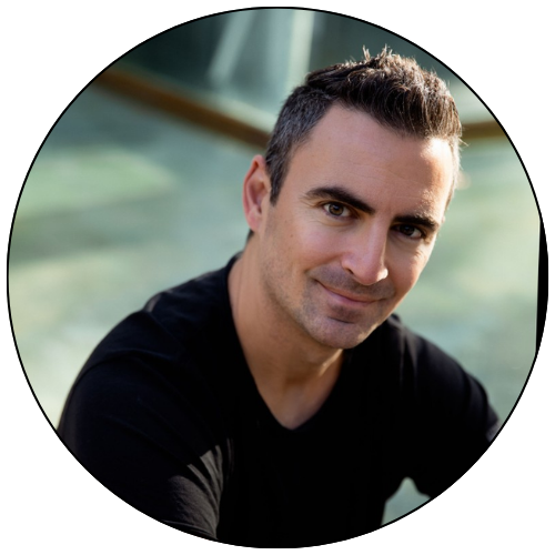 A young man with short dark hair and a slight smile, wearing a black shirt, looking into the camera with a blurred outdoor background.