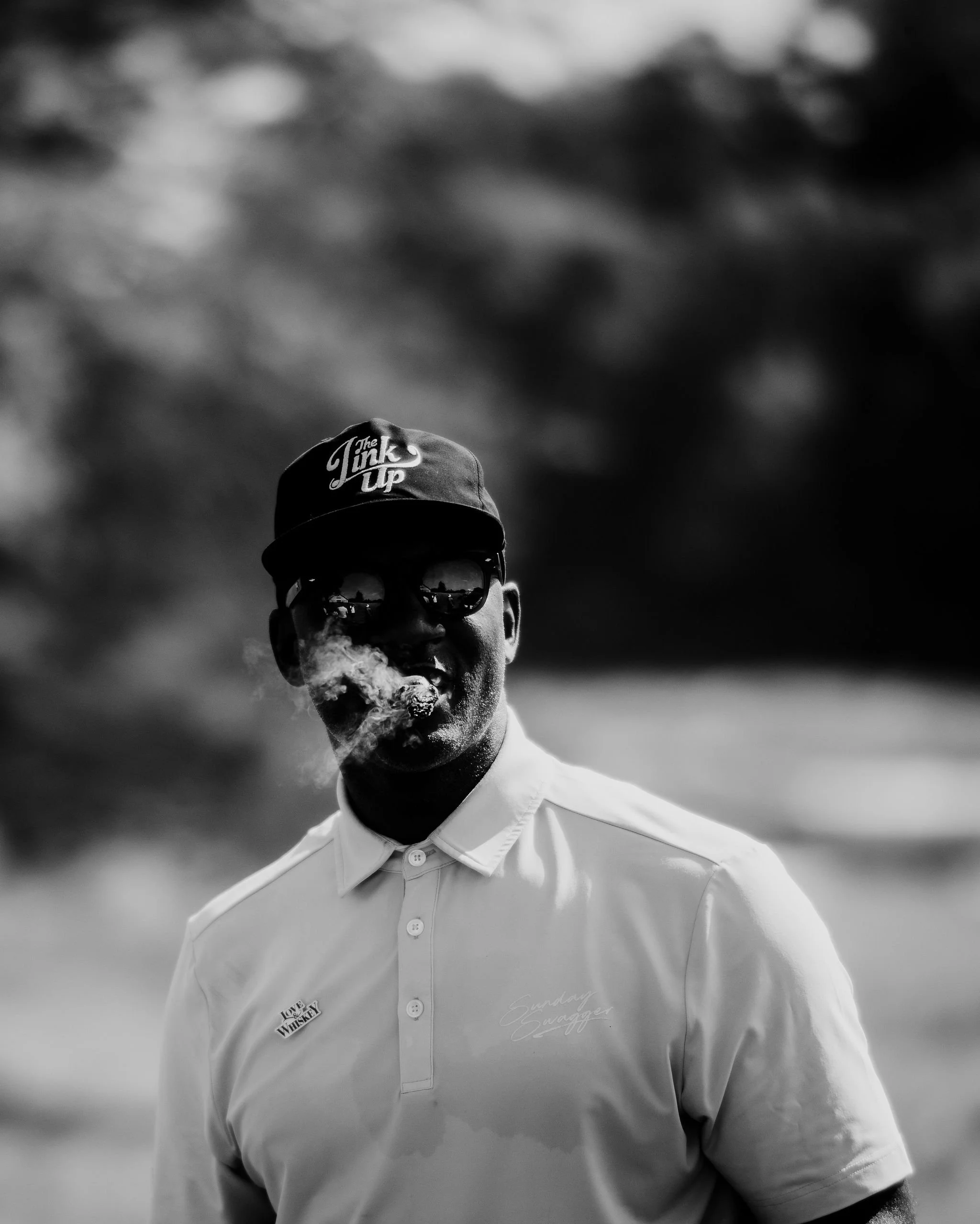 A man wearing sunglasses, a cap with the words "The Talk Up," and a collared shirt, smoking a cigar outdoors with a blurred natural background.