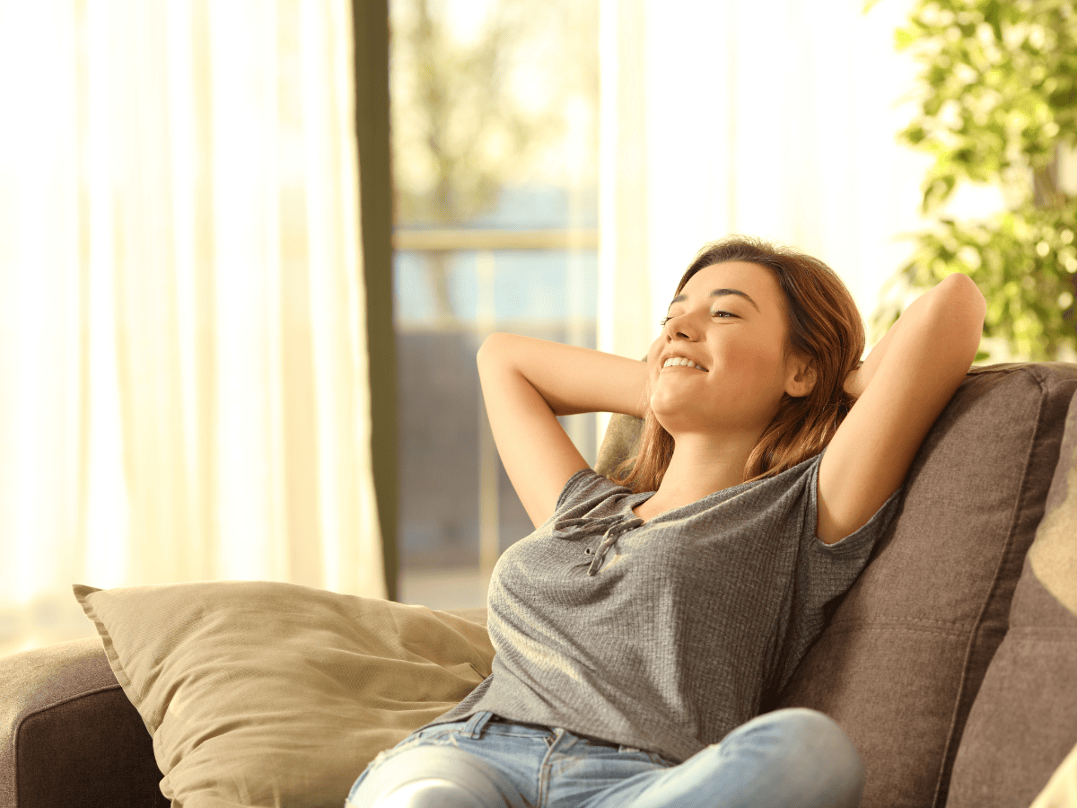 A woman is relaxing on a couch with her hands behind her head and smiling with her eyes closed, in a bright room with sunlight coming through the window.