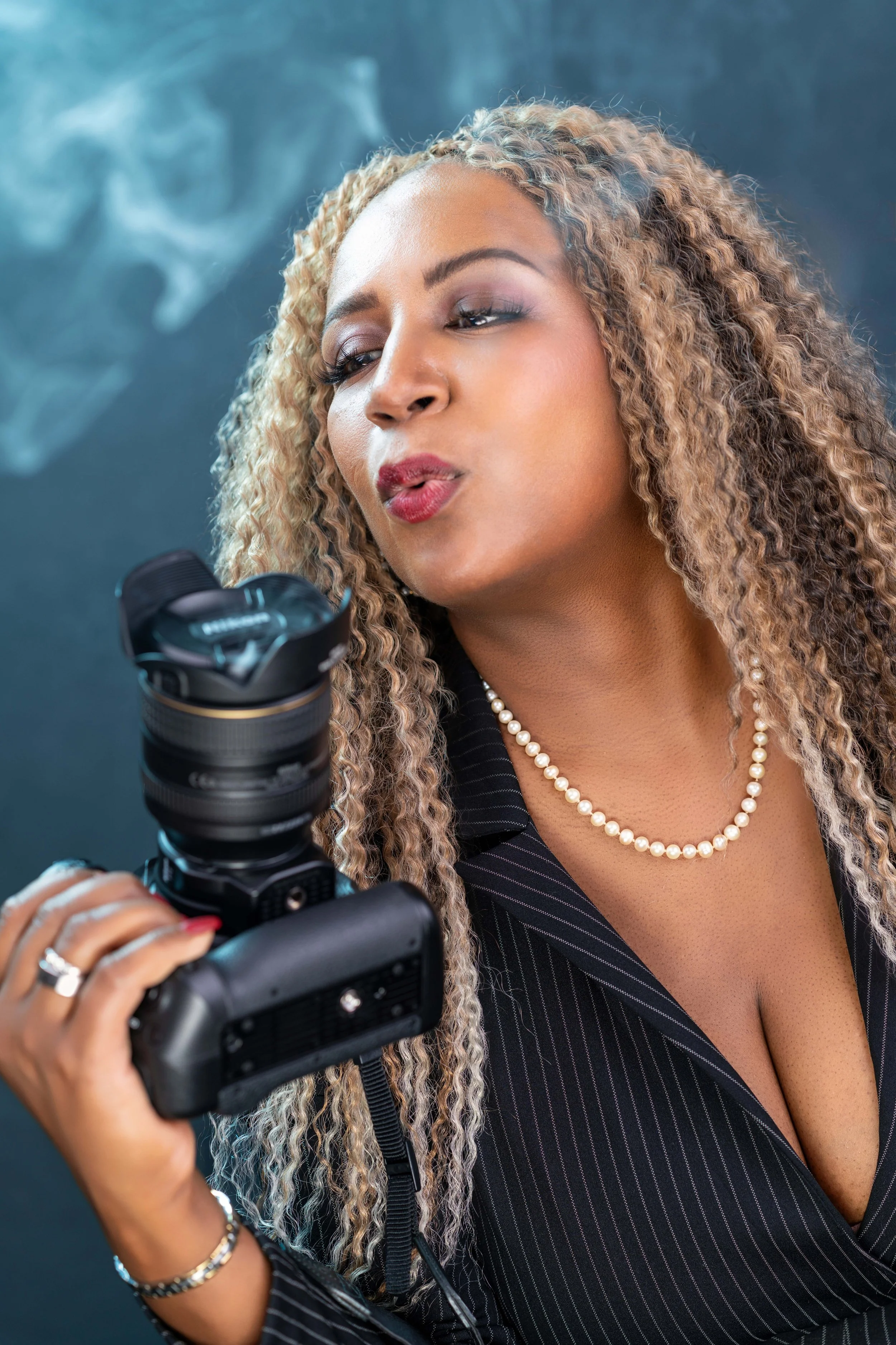 Portrait of Yevette, a female photographer with blonde curly hair and a pearl necklace, holding a professional DSLR camera in a black pinstripe jacket.