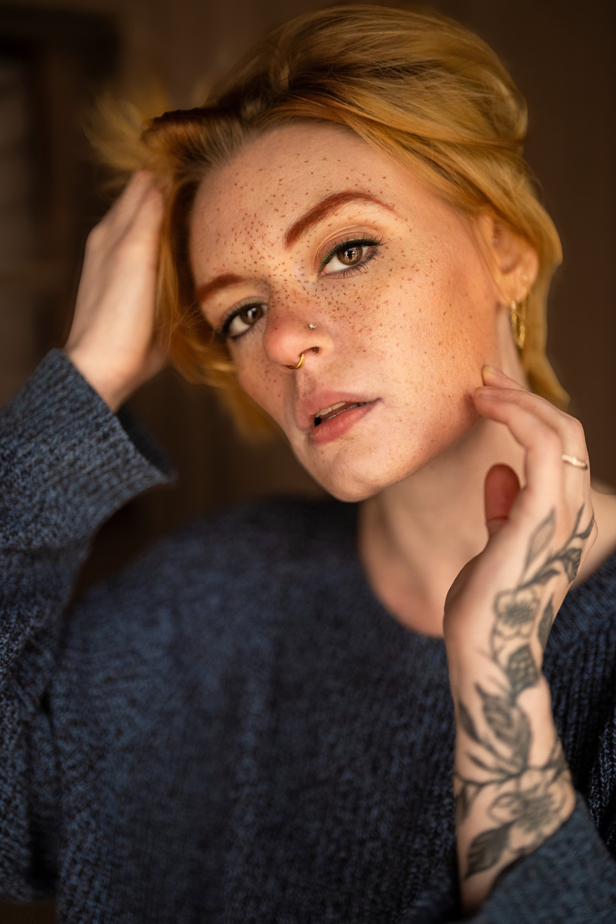 Close-up fashion portrait of Abigail, a young woman with short ginger hair, freckles, and a forearm tattoo, wearing a blue sweater.