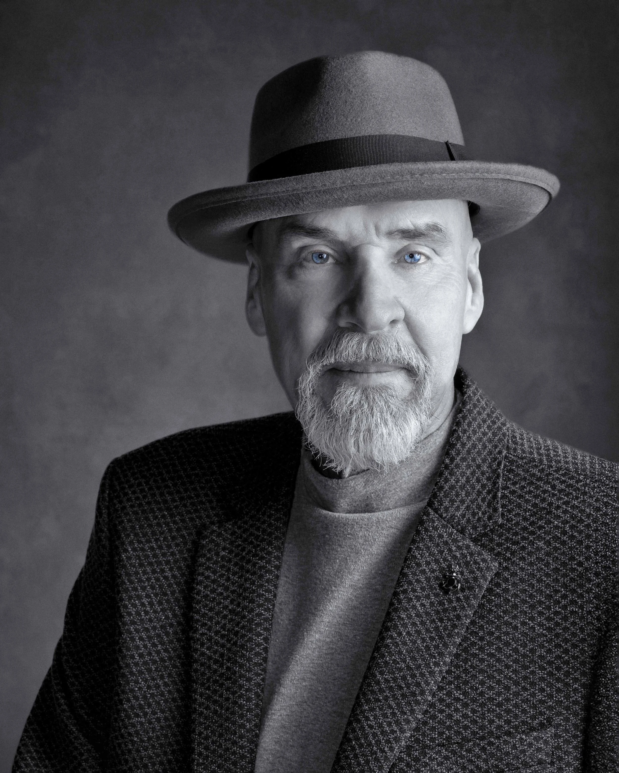 Black and white portrait of a confident man with a gray beard and fedora, featuring striking blue eyes.