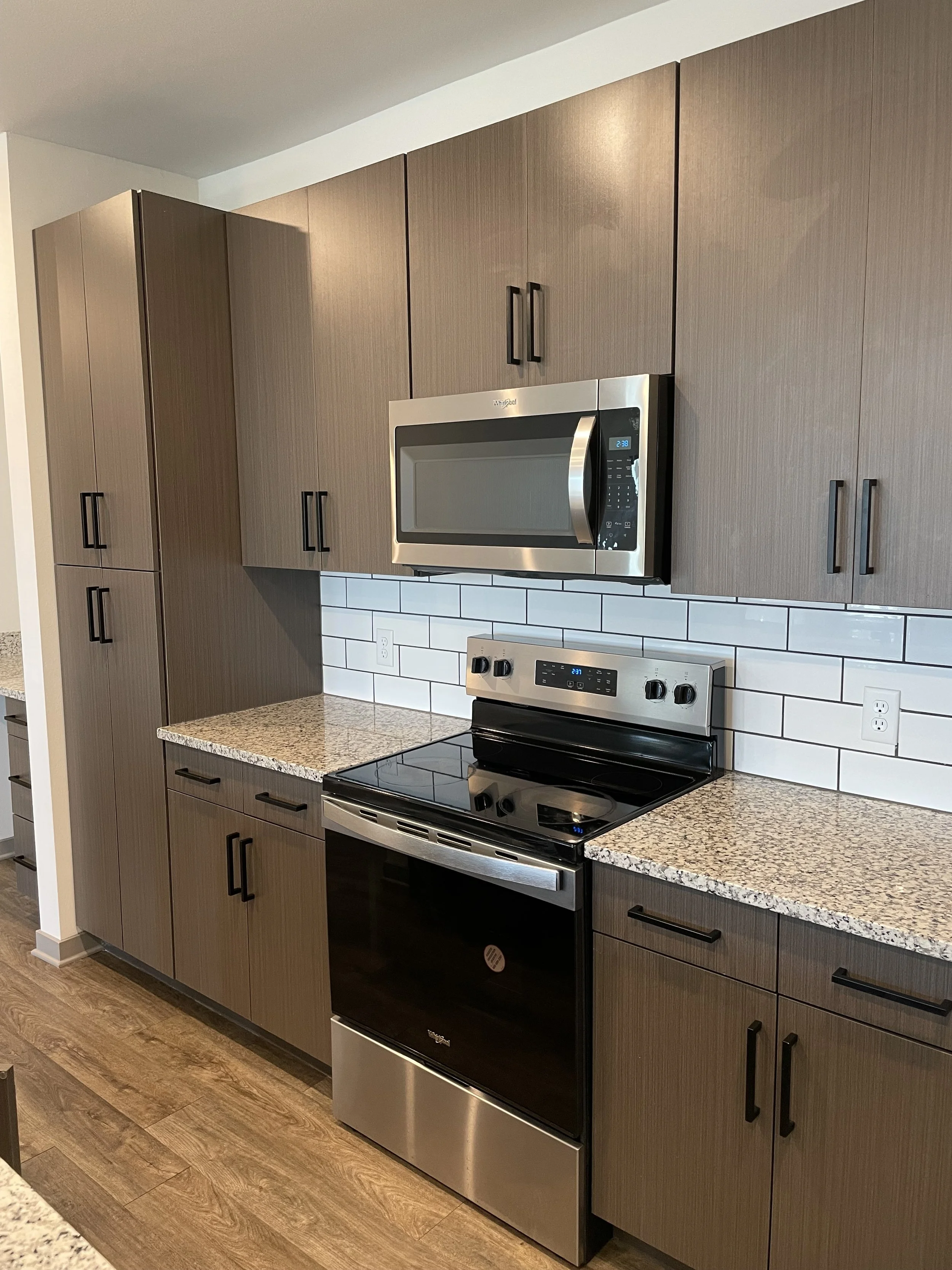 Kitchen with brown wood cabinets, granite countertops, a stainless steel microwave, and an electric stove with oven, white subway tile backsplash, and hardwood flooring.