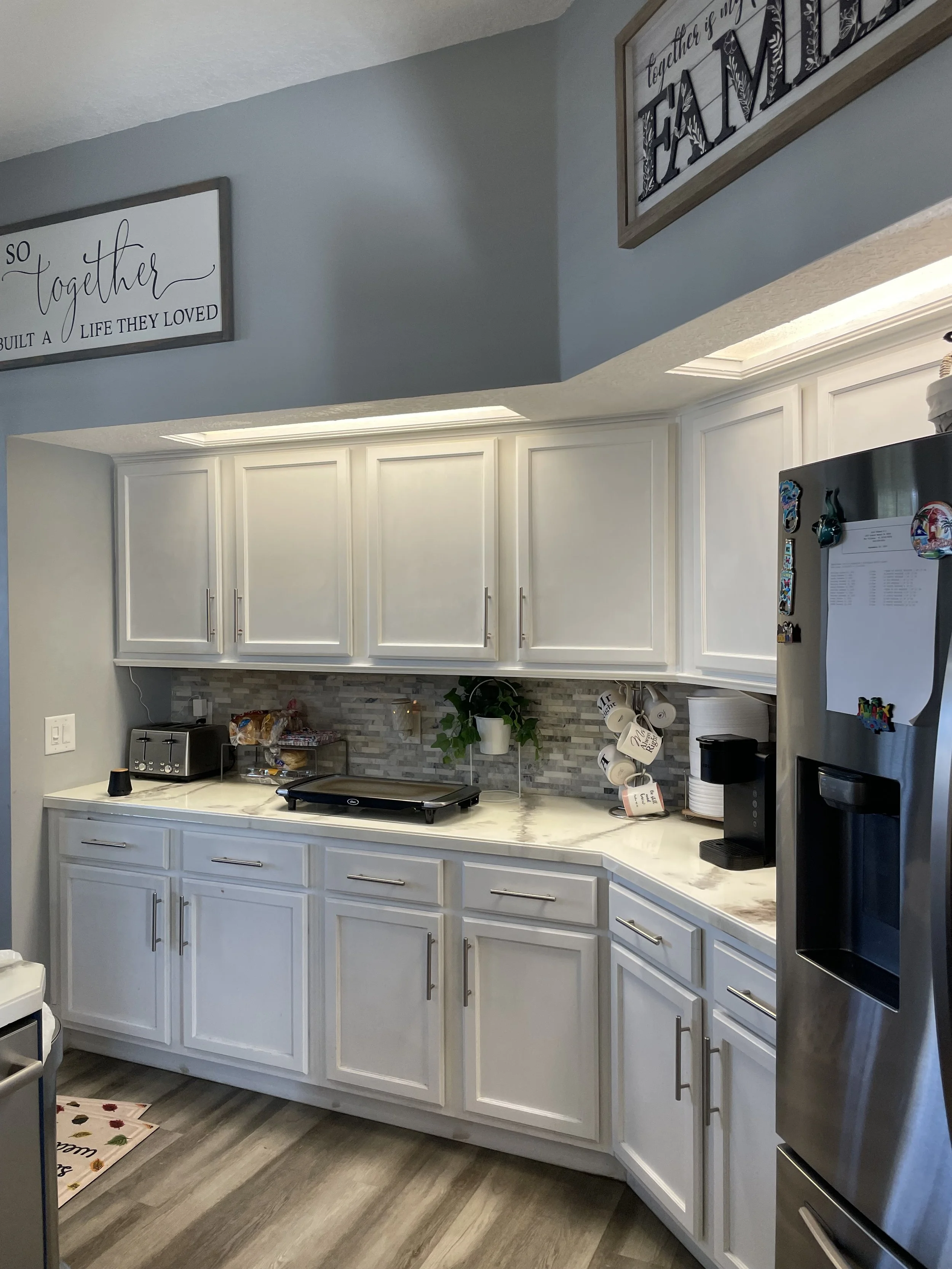Kitchen with white cabinets, a built-in microwave, a coffee maker on the right, a toaster, a hot plate, and a plant on the counter. There are decorative signs hanging on the wall above the cabinets.