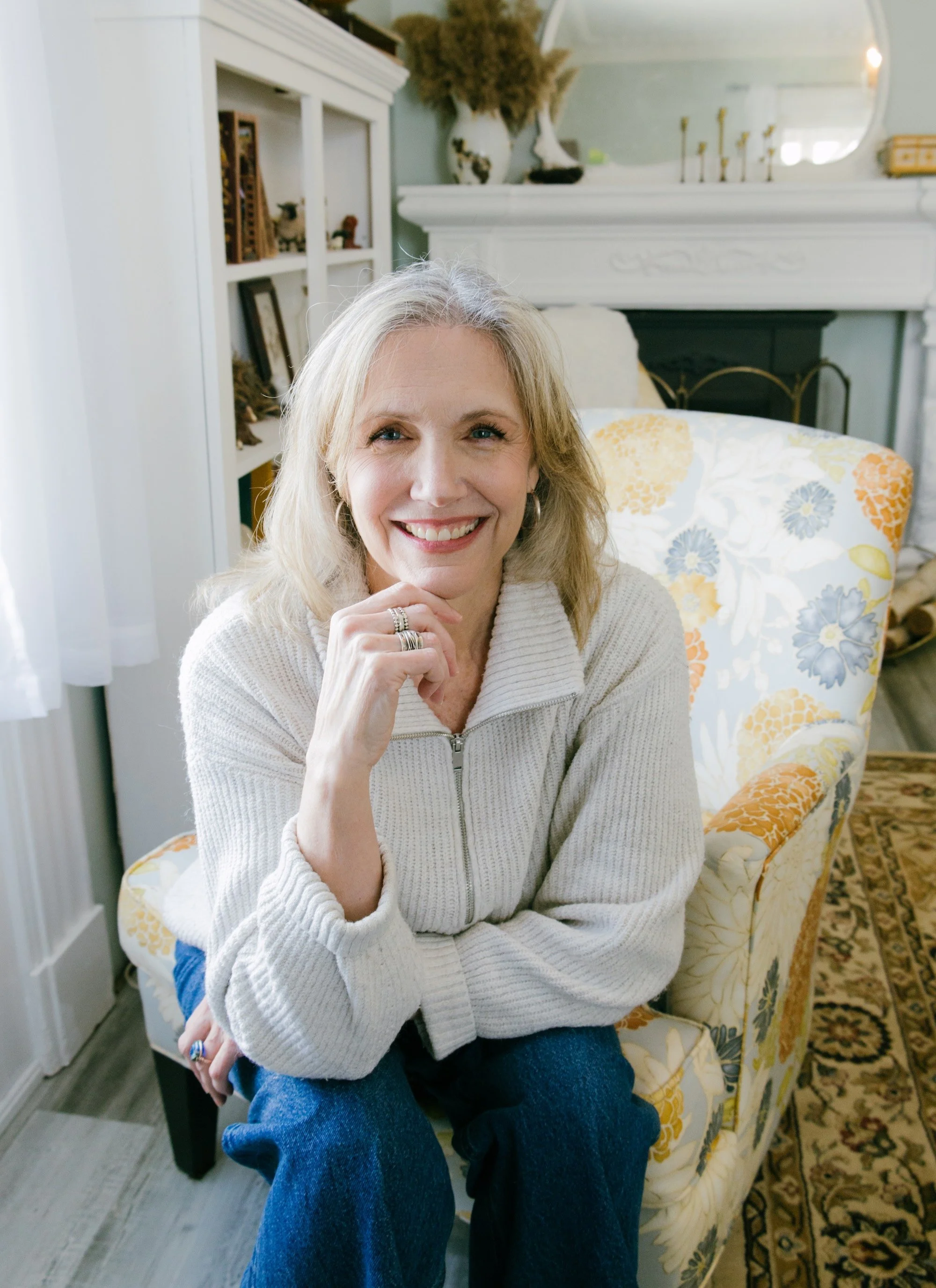A smiling woman with blonde hair and blue eyes sitting on a floral upholstered chair in a cozy, decorated room with a mirror, white fireplace, and bookshelf.