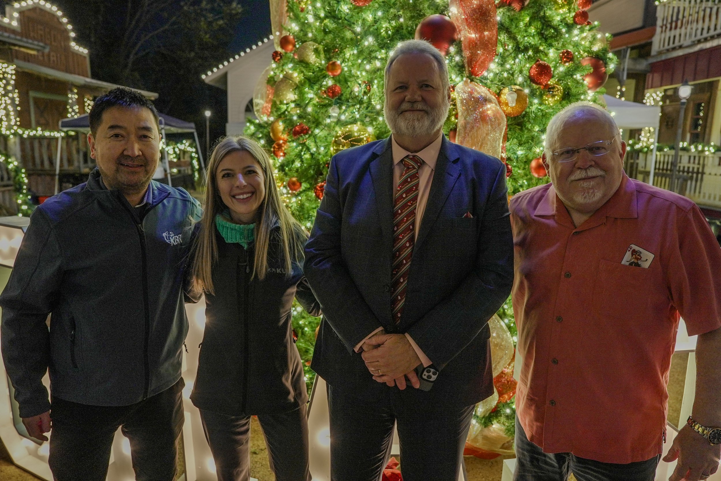 Four people posing in front of a decorated Christmas tree with lights and ornaments, during a nighttime outdoor event.