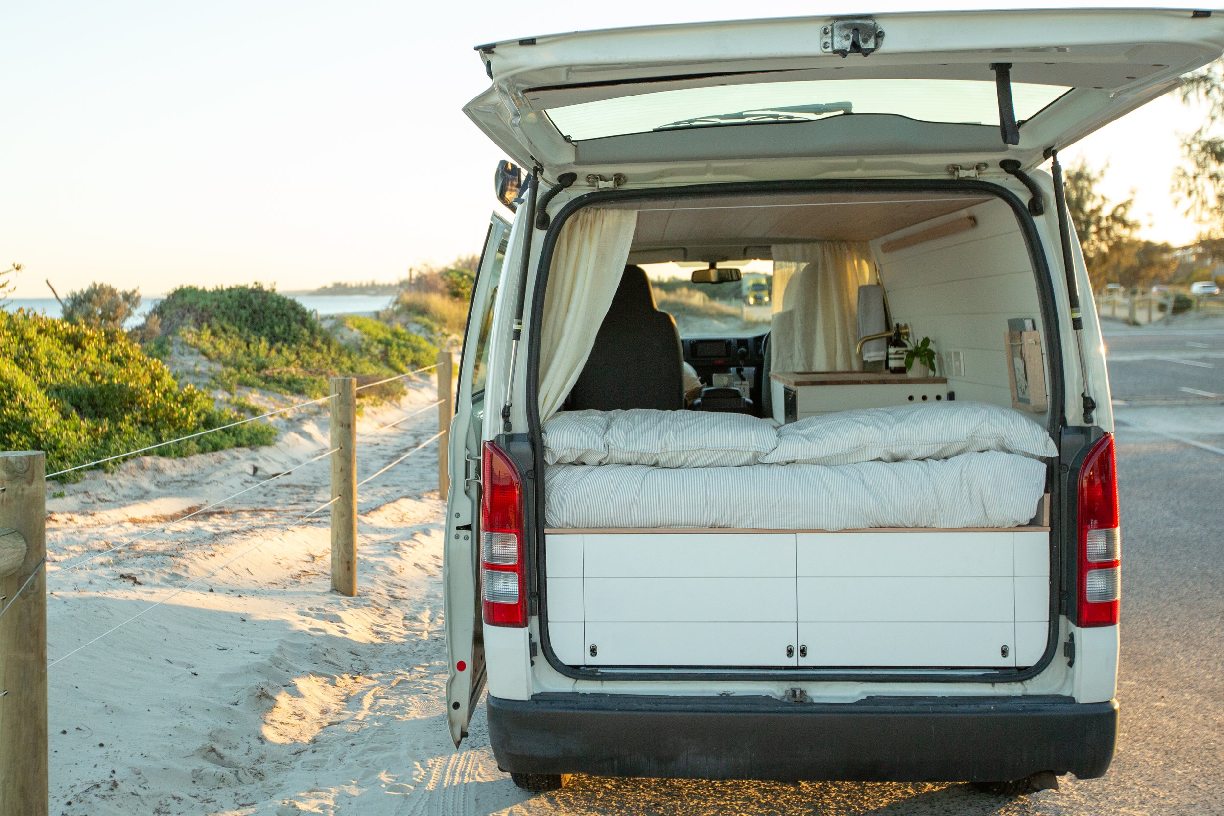 White camper van with open rear doors, showing an interior bed setup, parked near a sandy beach with greenery.