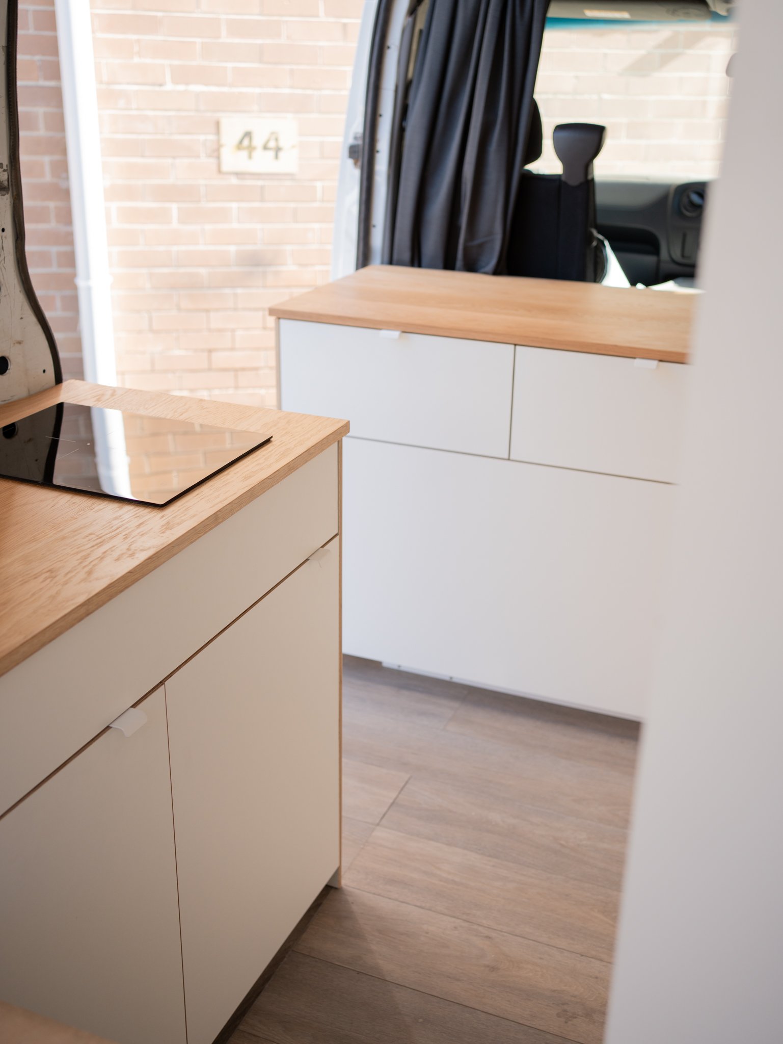 Interior view of a modern kitchen with cream cabinets, a wooden countertop, a black cooktop, and a view into the driver's area of a vehicle through an open door.