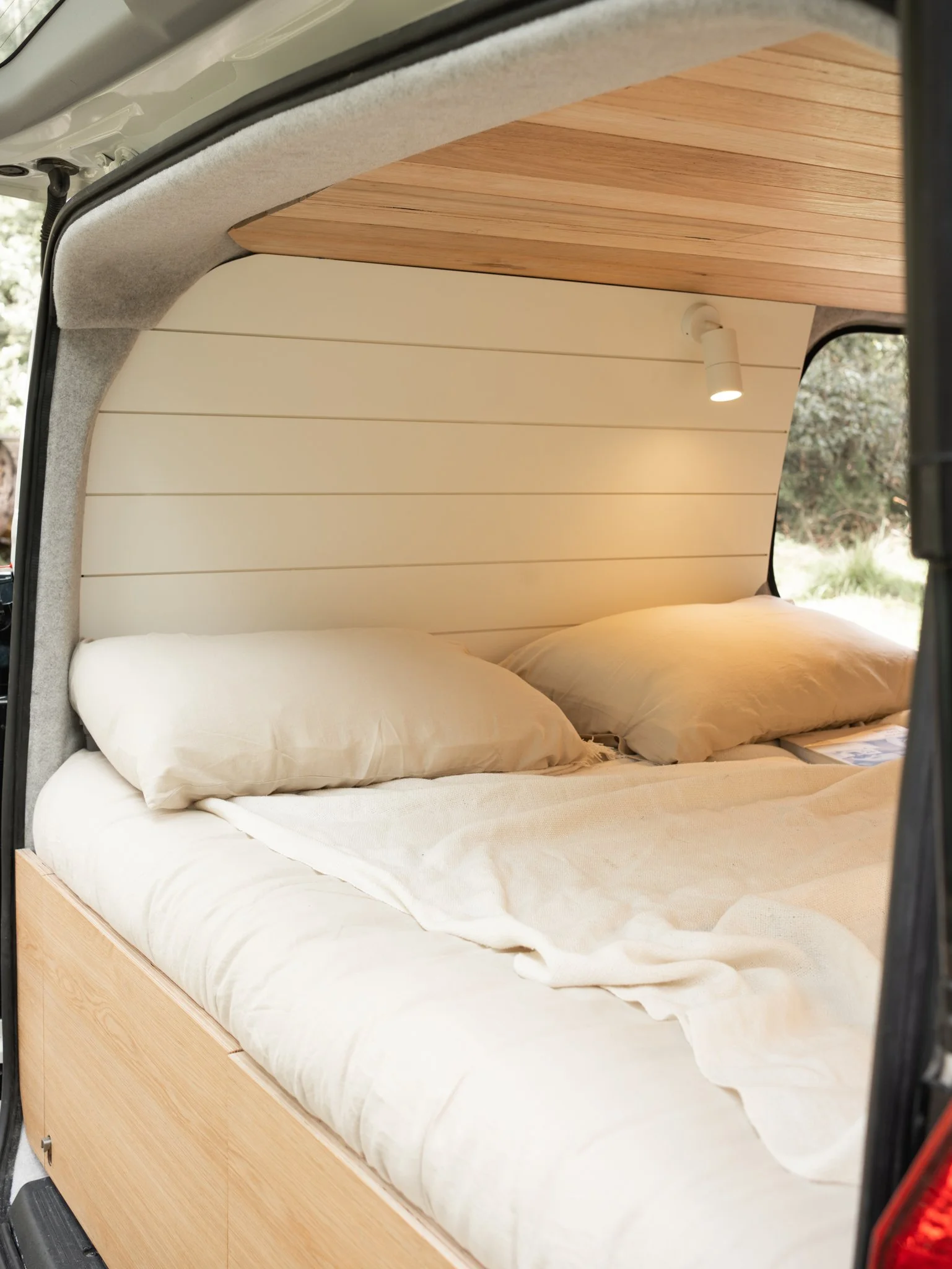 Interior of a camper van with a neatly made bed featuring beige pillows and blankets, wood paneling on the ceiling, and a small adjustable reading light.