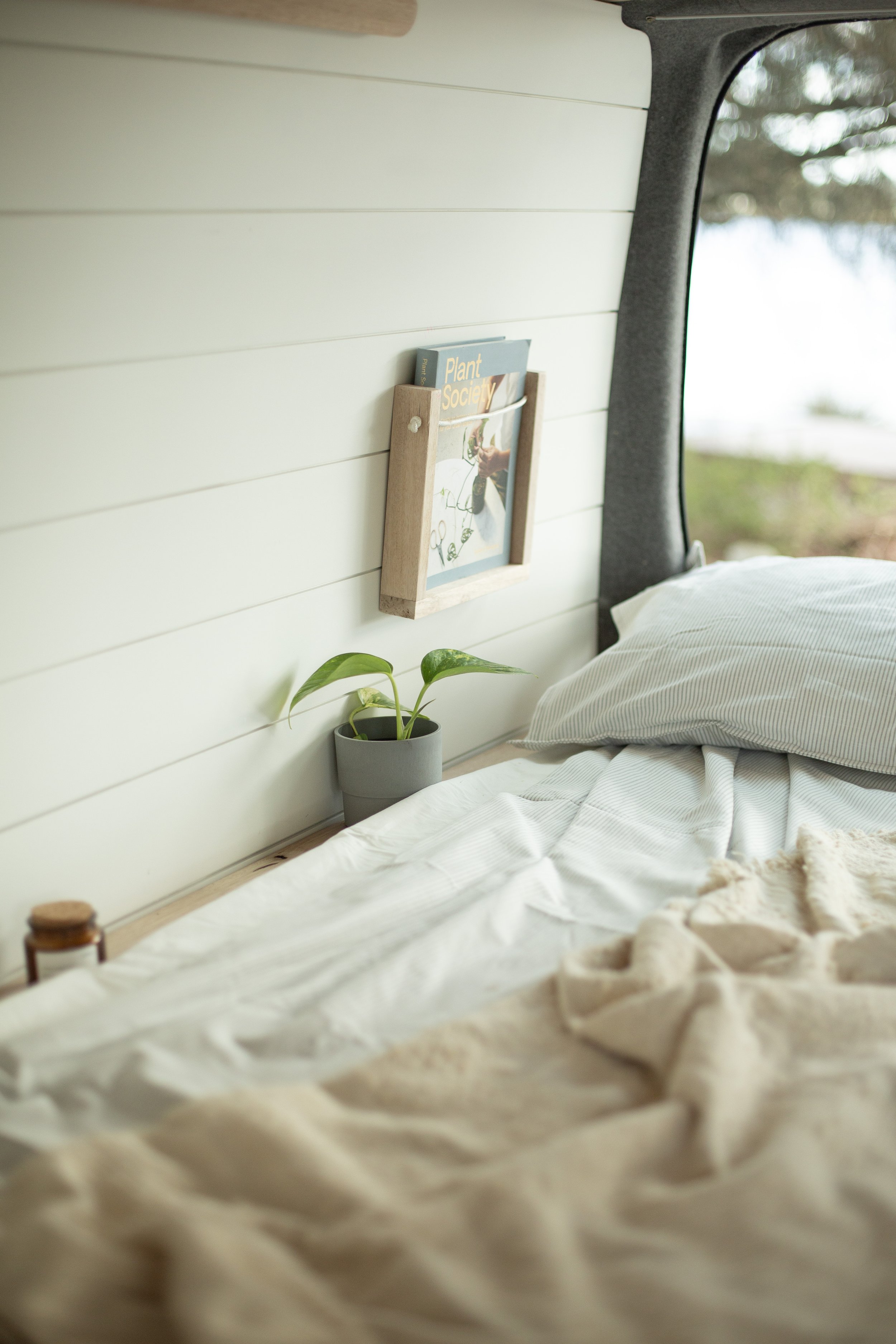 Interior of a cozy van with white shiplap walls, a small potted plant on a shelf, a book titled 'Plant Society' in a wooden holder, and a bed with light bedding next to a window.