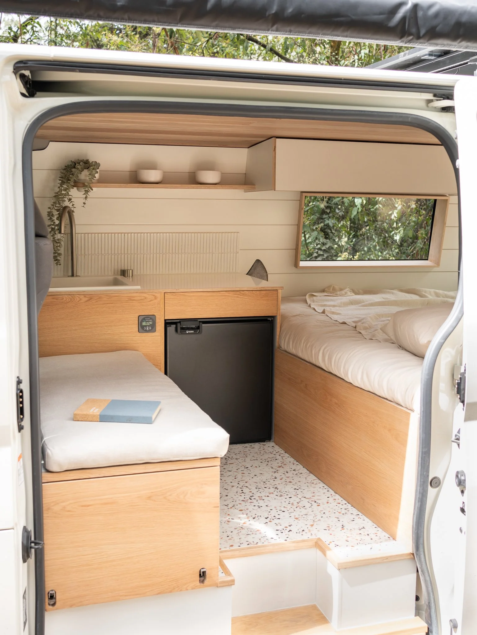 Interior of a small camper van with wooden furnishings, a bed, a small refrigerator, a kitchenette with a sink, and a window showing greenery outside.