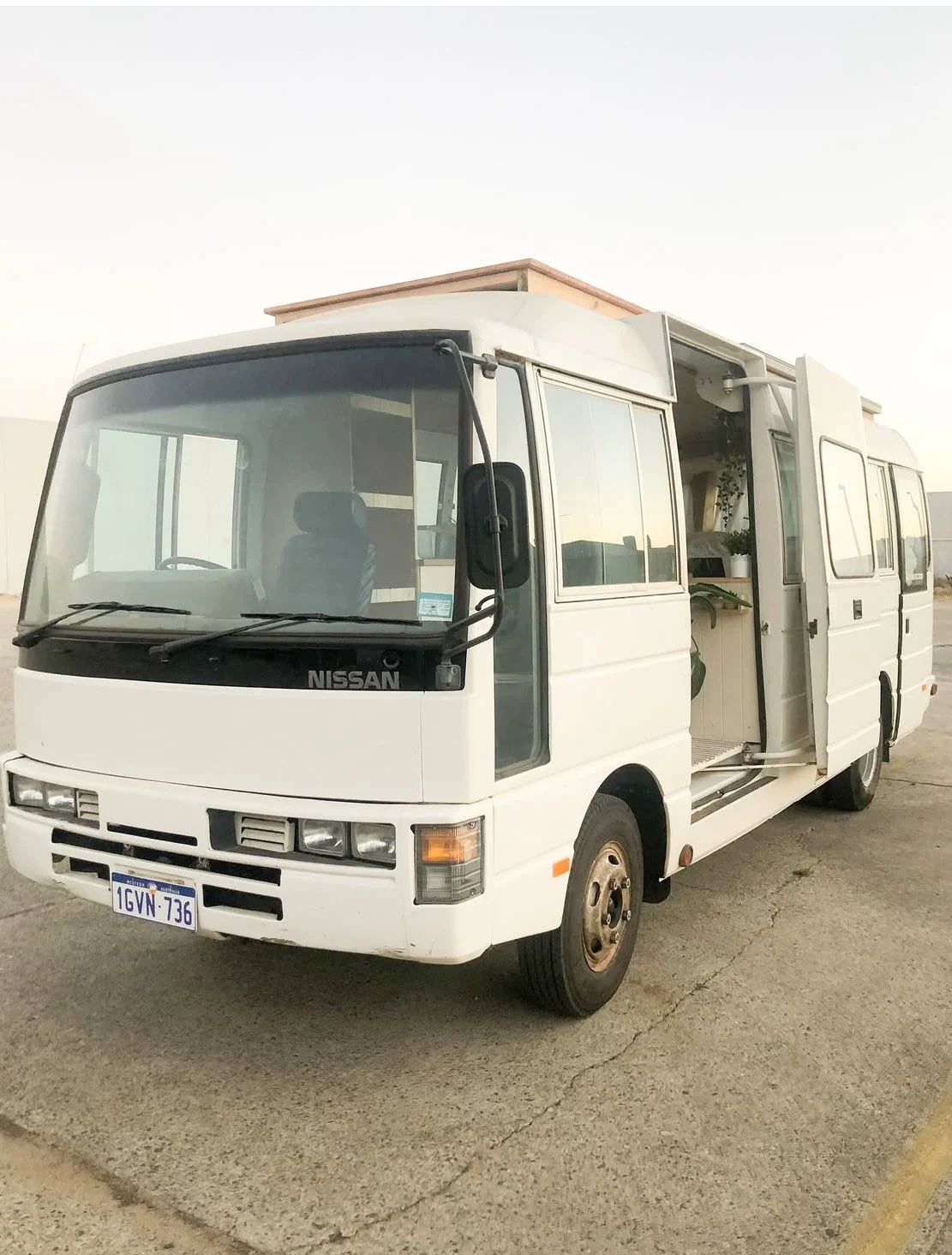 A white vintage Nissan camper van with its side door open, showing indoor plants inside, parked on a paved surface under a clear sky.