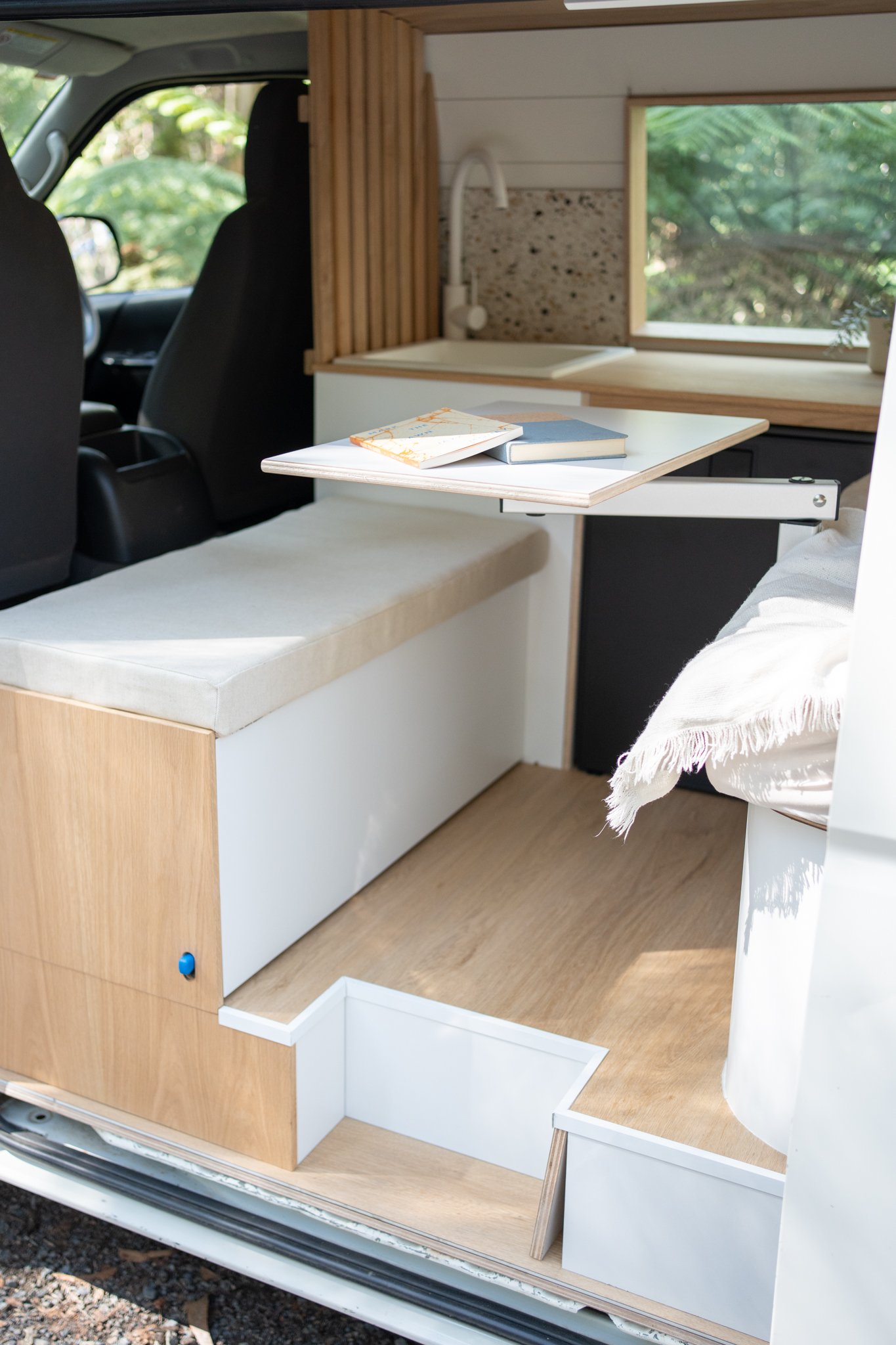 Interior of a camper van with a bench, folding table, books, sink, and window.