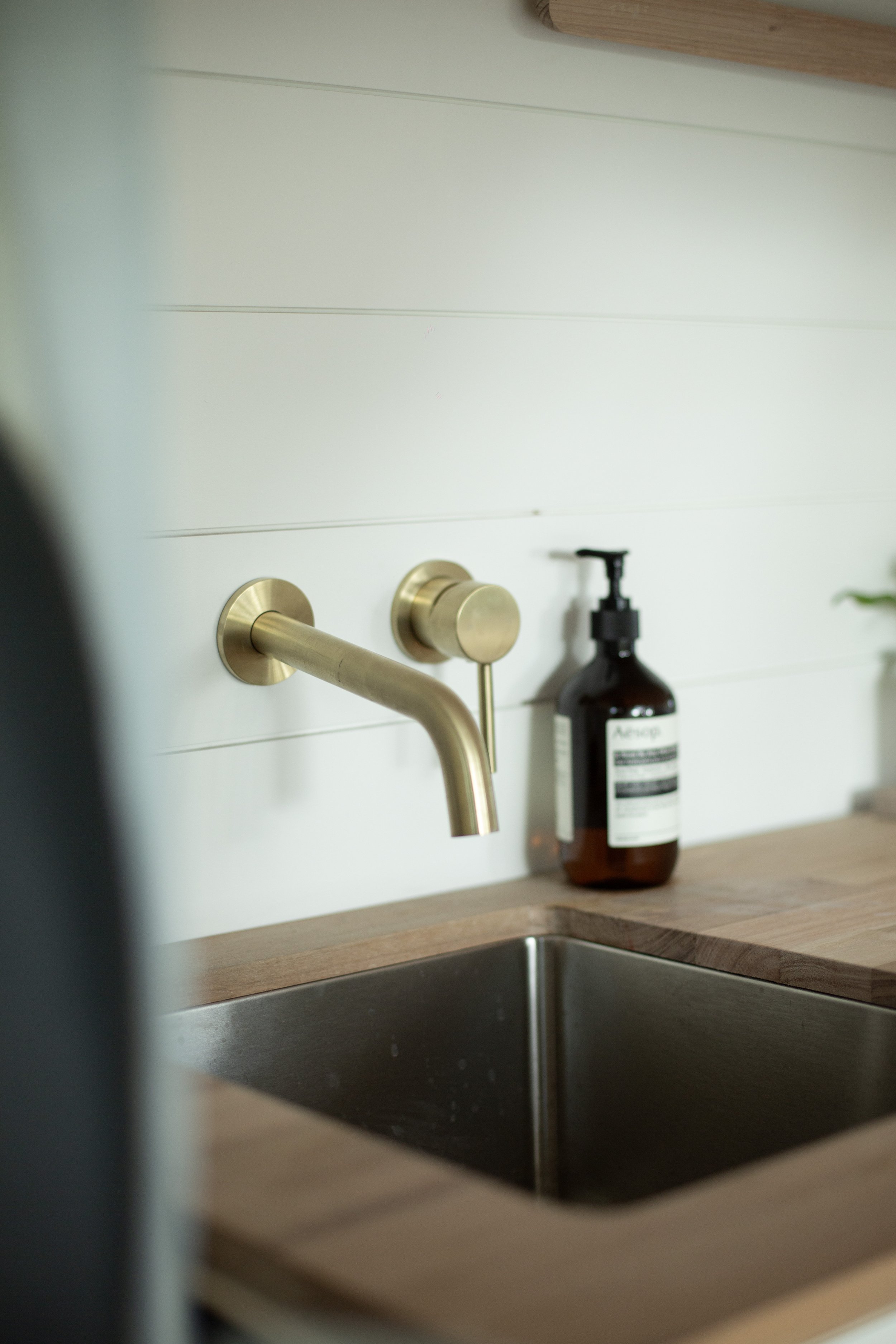 Modern kitchen sink with brass wall-mounted faucet and wooden countertop, bottle of soap on the side.