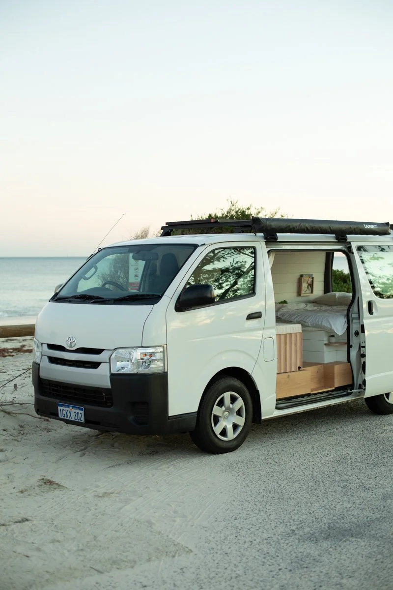 A white camper van parked on a beach near the ocean, with the sliding door open revealing a bed inside.