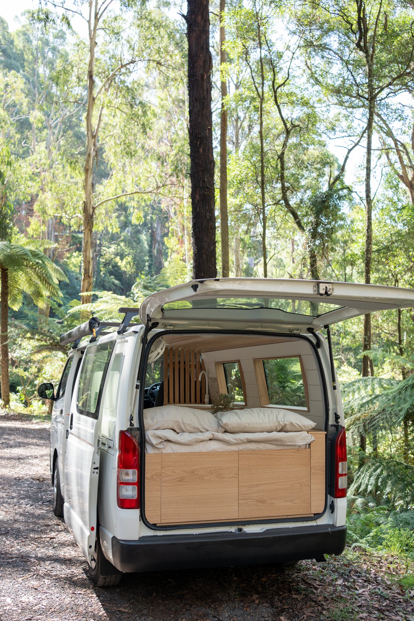 A white camper van with its back door open, revealing a cozy interior setup with a bed, parked in a lush forest.