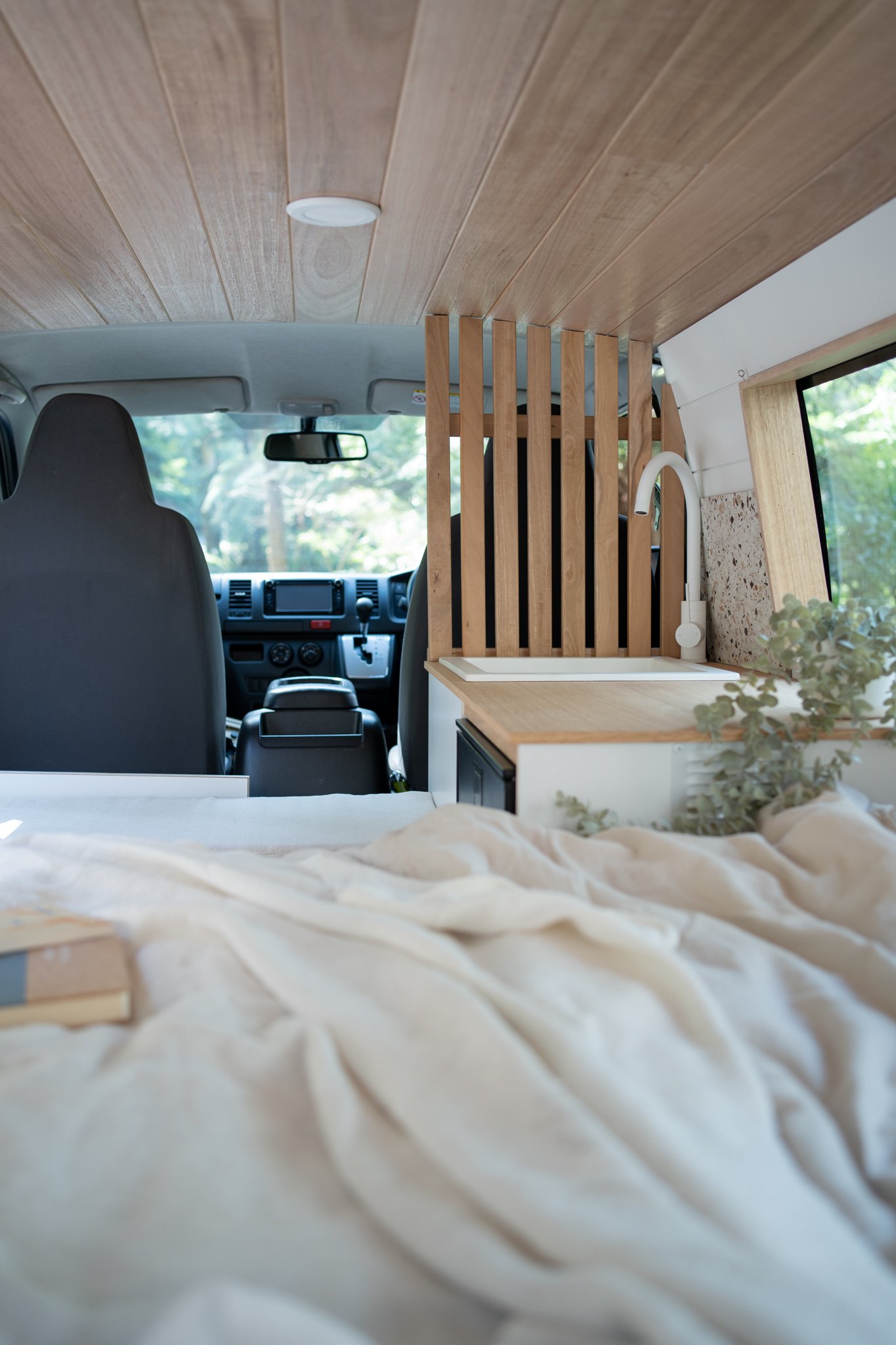 Interior of a camper van featuring a wooden ceiling, a cozy bed with a light blanket and a book, a small countertop with a sink, and a wooden partition.