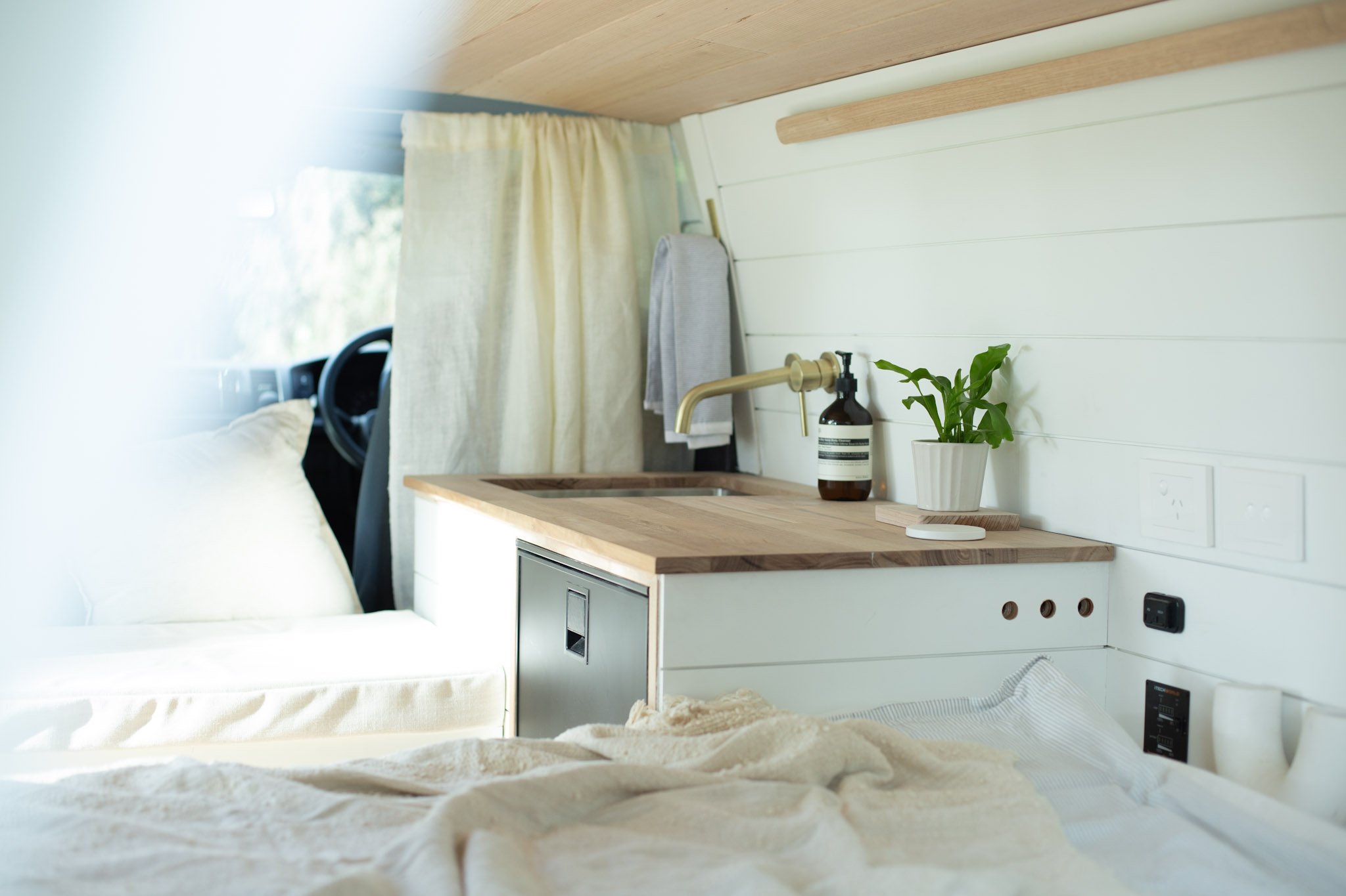 Interior of a camper van with a kitchenette featuring a wooden countertop, sink, faucet, potted plant, and soap dispenser. Light-colored curtains and bedding are also visible.