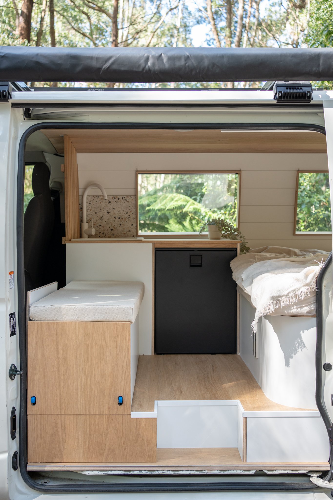 Interior of a camper van with wooden flooring, a compact kitchen, and a bed. The space includes a sink, counter, and a small refrigerator, highlighted by windows with wooded views.