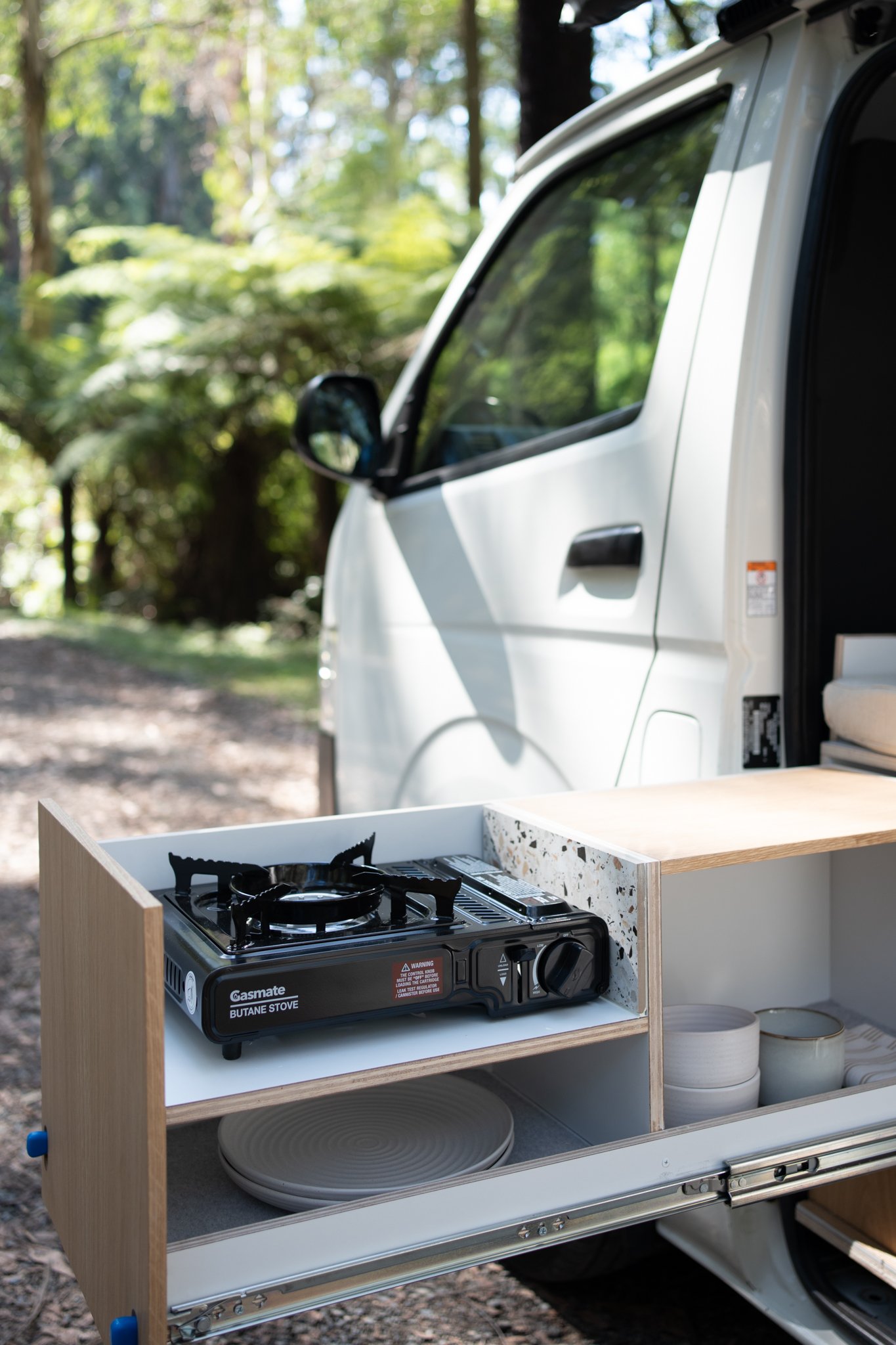 Van with an outdoor kitchen setup, featuring a pull-out wooden drawer holding a portable butane stove and kitchenware, set in a forested area.