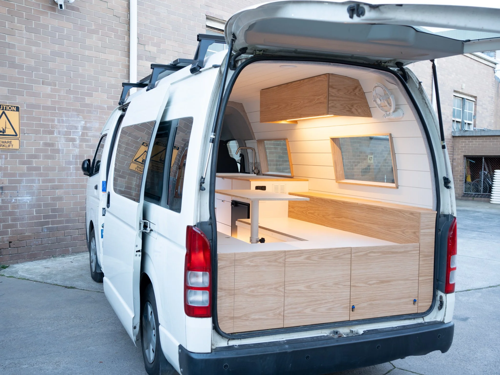 Interior of a white van converted into a tiny mobile workspace with wooden cabinetry, windows, a small sink, and a counter with a person working inside.