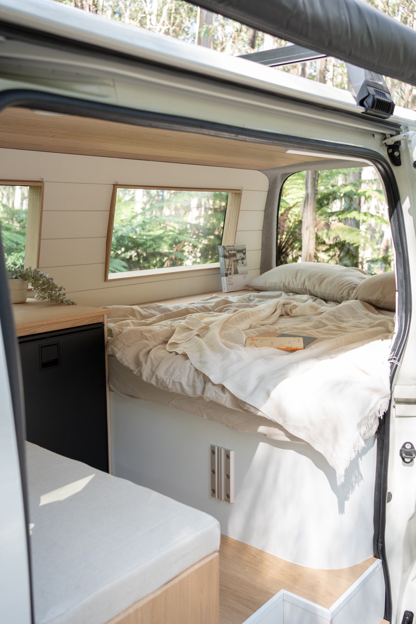 Interior of a camper van with a bed, cream-colored bedding, windows with views of trees, and a small cabinet.