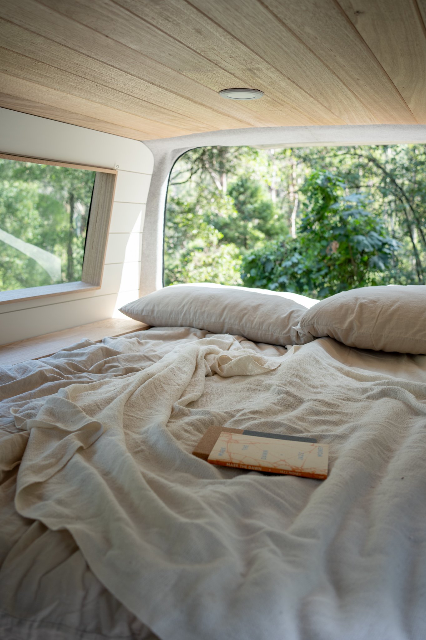 Cozy bed with beige linens inside a camper van, surrounded by lush greenery visible through a large window.
