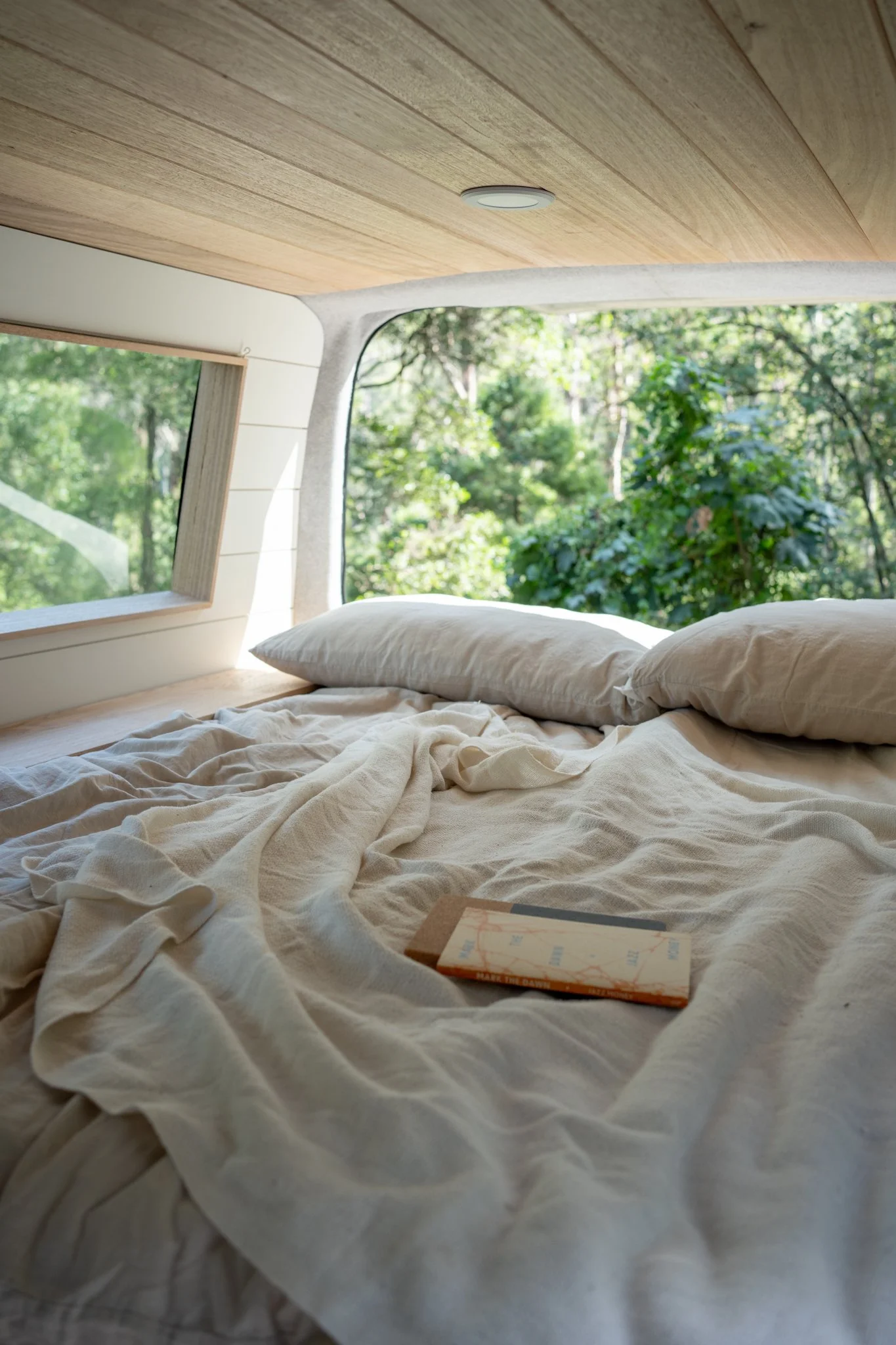 A cozy bed with cream-colored sheets and pillows inside a camper van with large windows showing green trees outside.