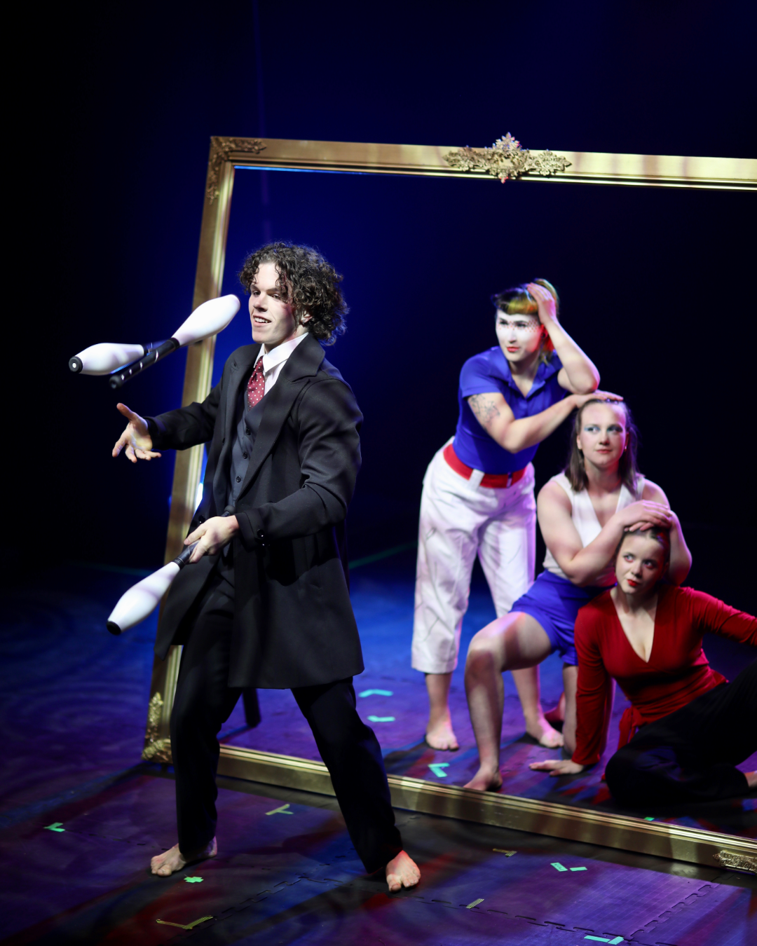 Performers in theatrical costumes on stage, with a large gold frame in the background, during a theatrical scene.