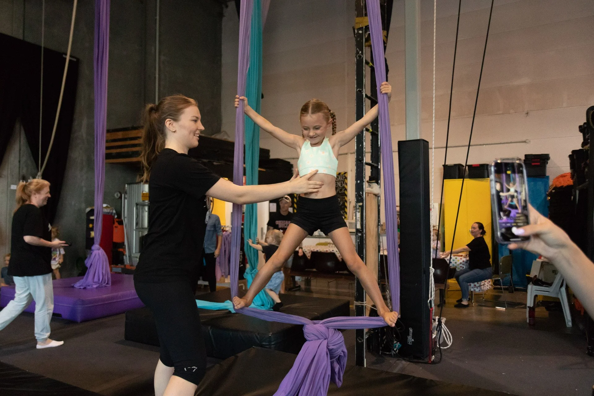 A young girl in a white tank top and black shorts practicing aerial silks with assistance in a gym. She is smiling, holding her arms out, with her legs spread wide, and is supported by a woman in black. Other children and adults are visible in the background.