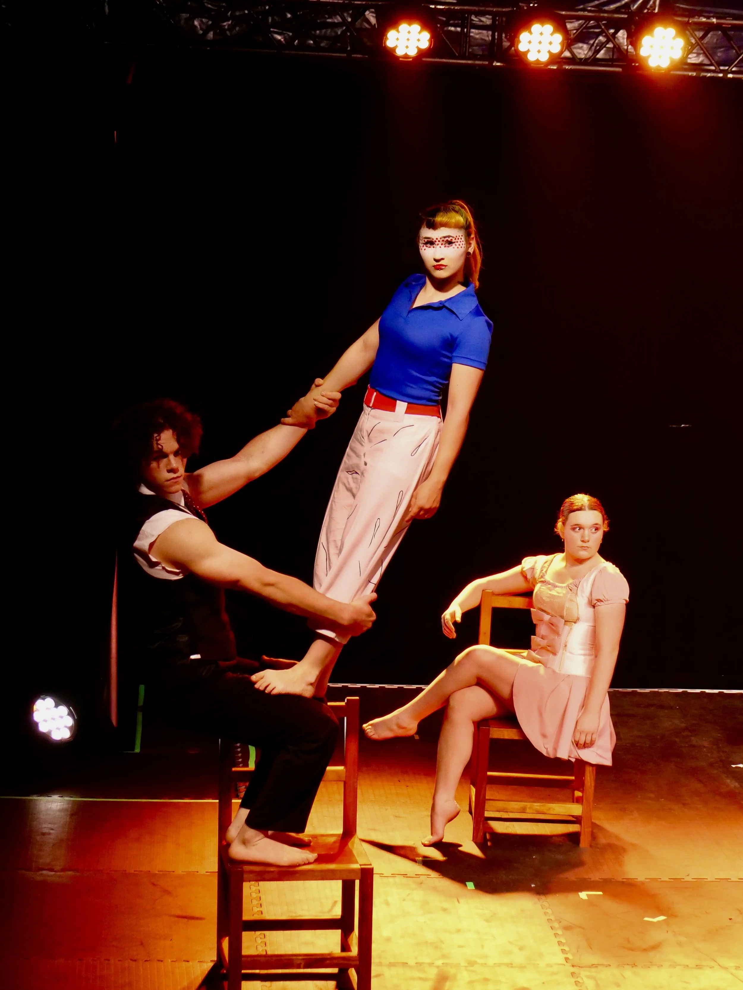 Three performers on stage, two seated on chairs and one standing, holding the other's hand, under stage lights.