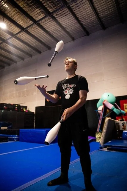 A man in black clothing juggles three white clubs in an indoor gym with a blue mat, industrial ceiling, and padded walls.