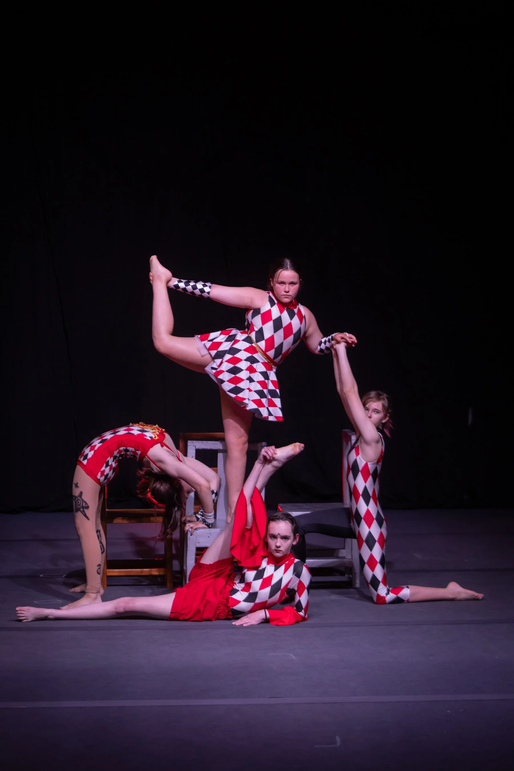Four female performers in harlequin costumes executing a balance routine on stage with a black background.