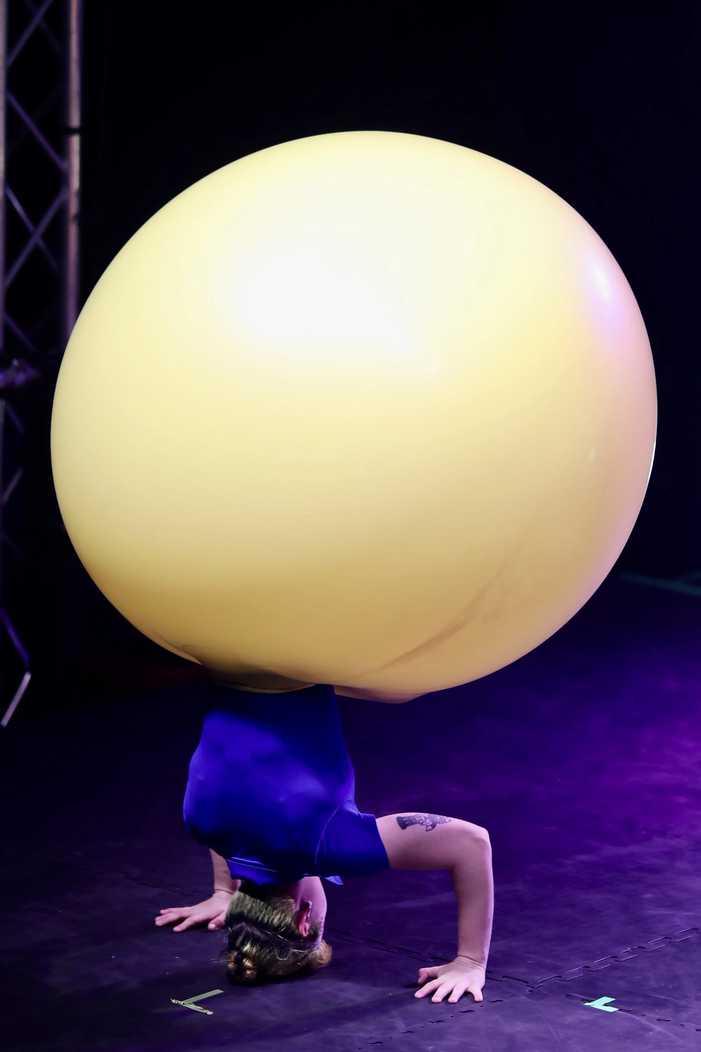 A performer balancing on hands in a handstand with a large glowing yellow sphere on their back during a performance.