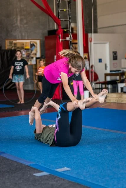A young girl in a pink shirt practicing gymnastics with an instructor lying on the blue mat, holding her feet as she balances.