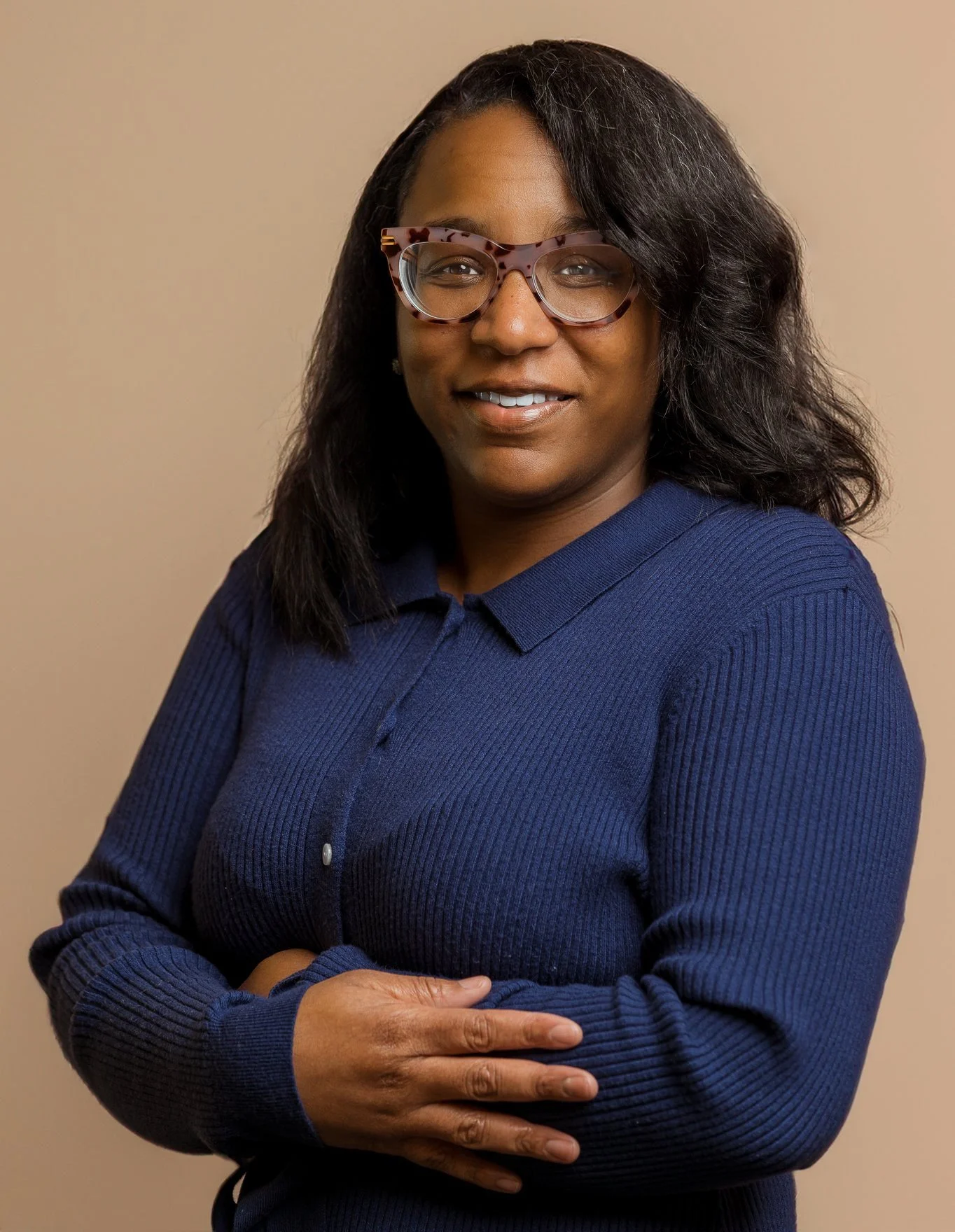 A smiling African American woman with black wavy hair wearing large tortoiseshell glasses and a dark blue ribbed button-up shirt posing with arms crossed against a beige background.