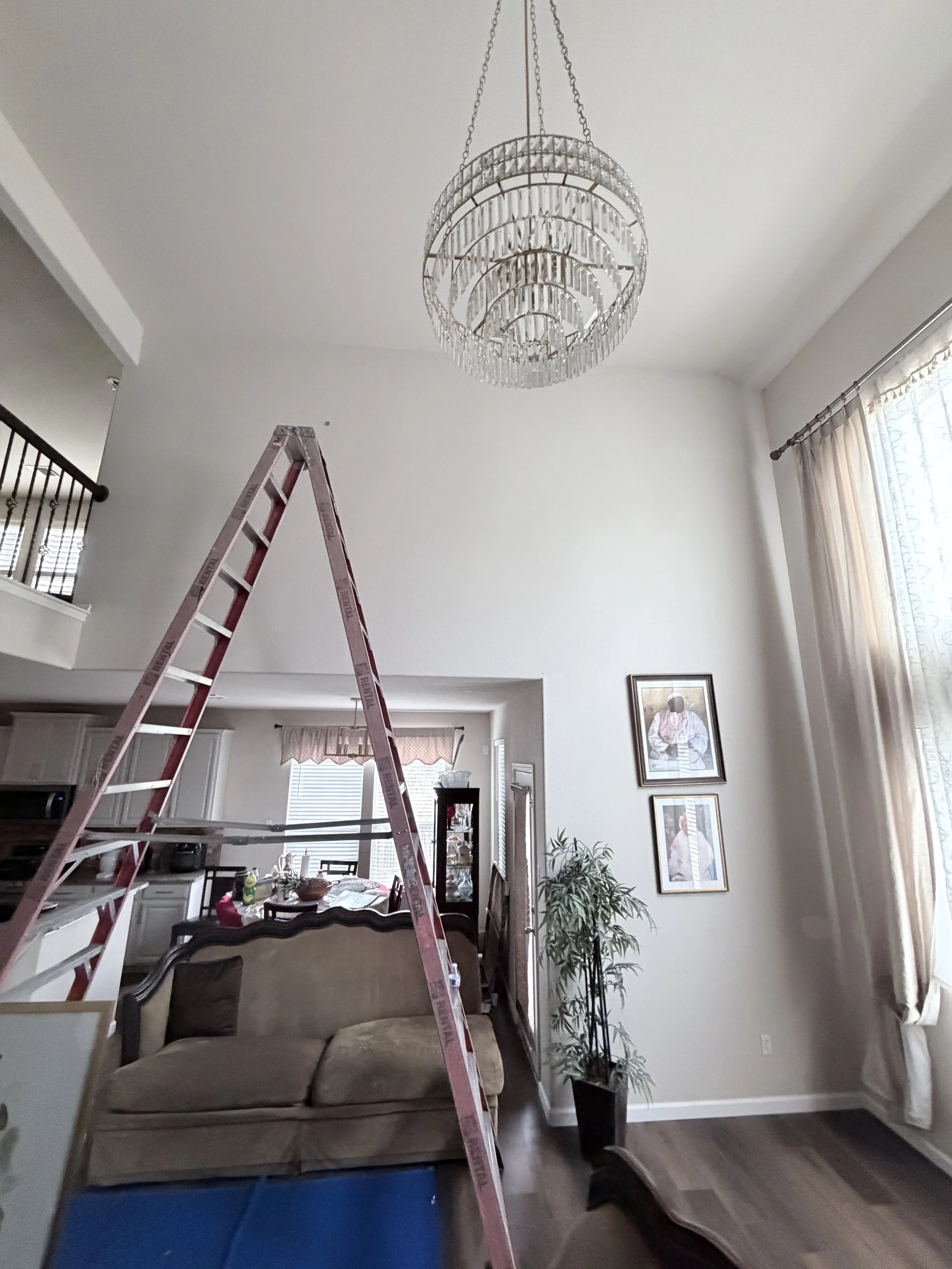 Chandelier installed in high ceiling living room using proper ladder setup and secure mounting.