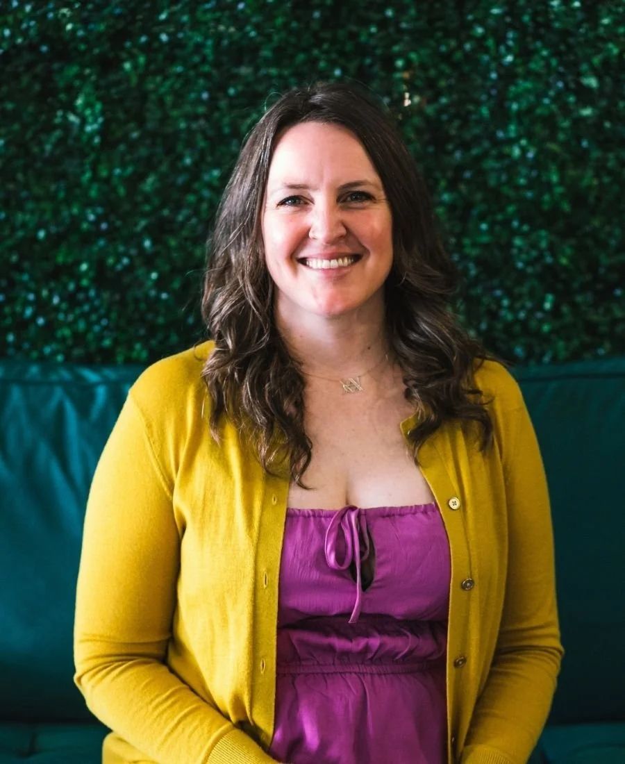 A woman with long, wavy dark hair, wearing a yellow cardigan over a purple dress, smiling at the camera with a green, textured background.