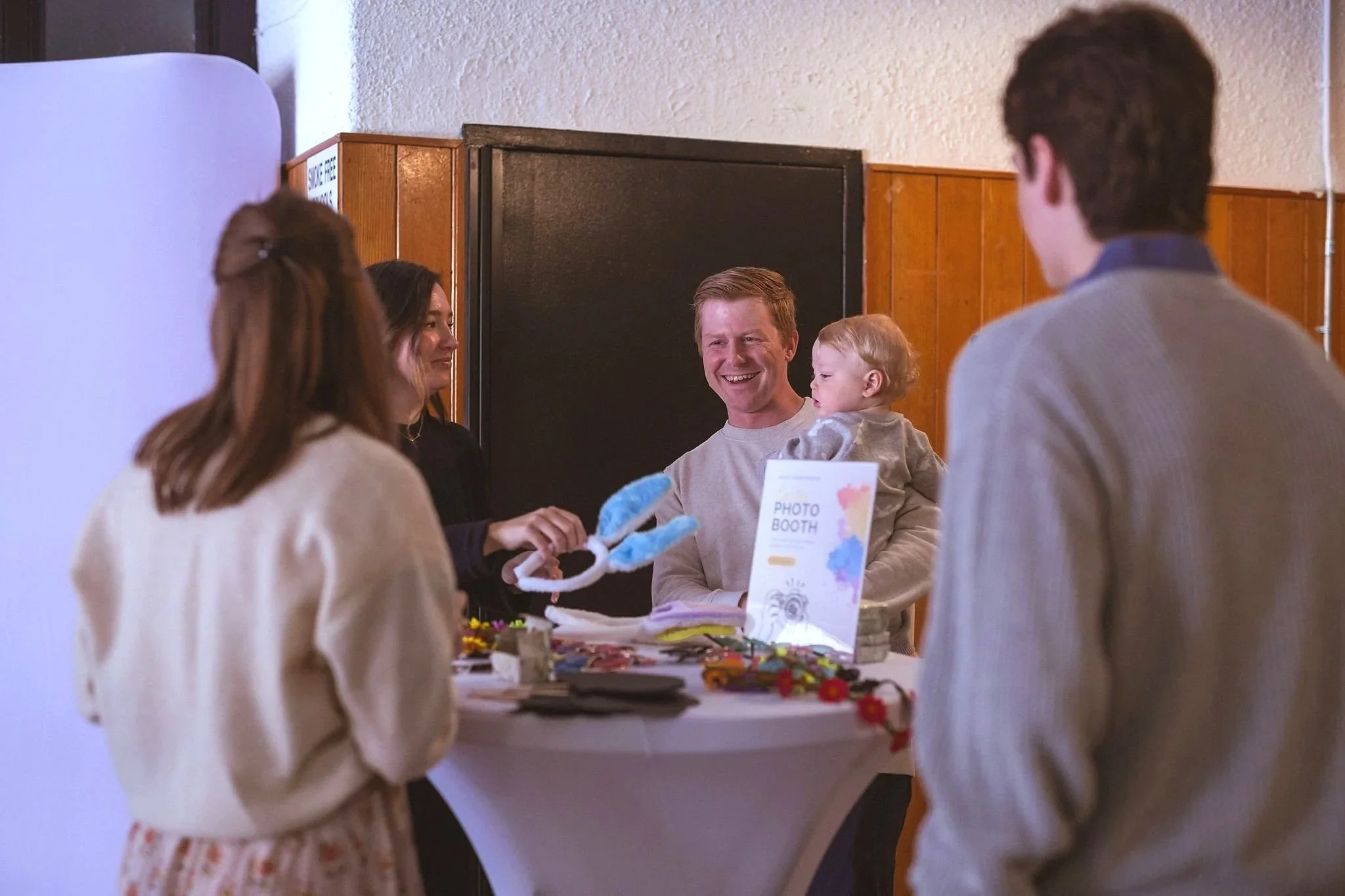 A group of five people, including a man holding a young child, gather around a photo booth at a social event in a room with wood-paneled walls, enjoying a moment of laughter and conversation.