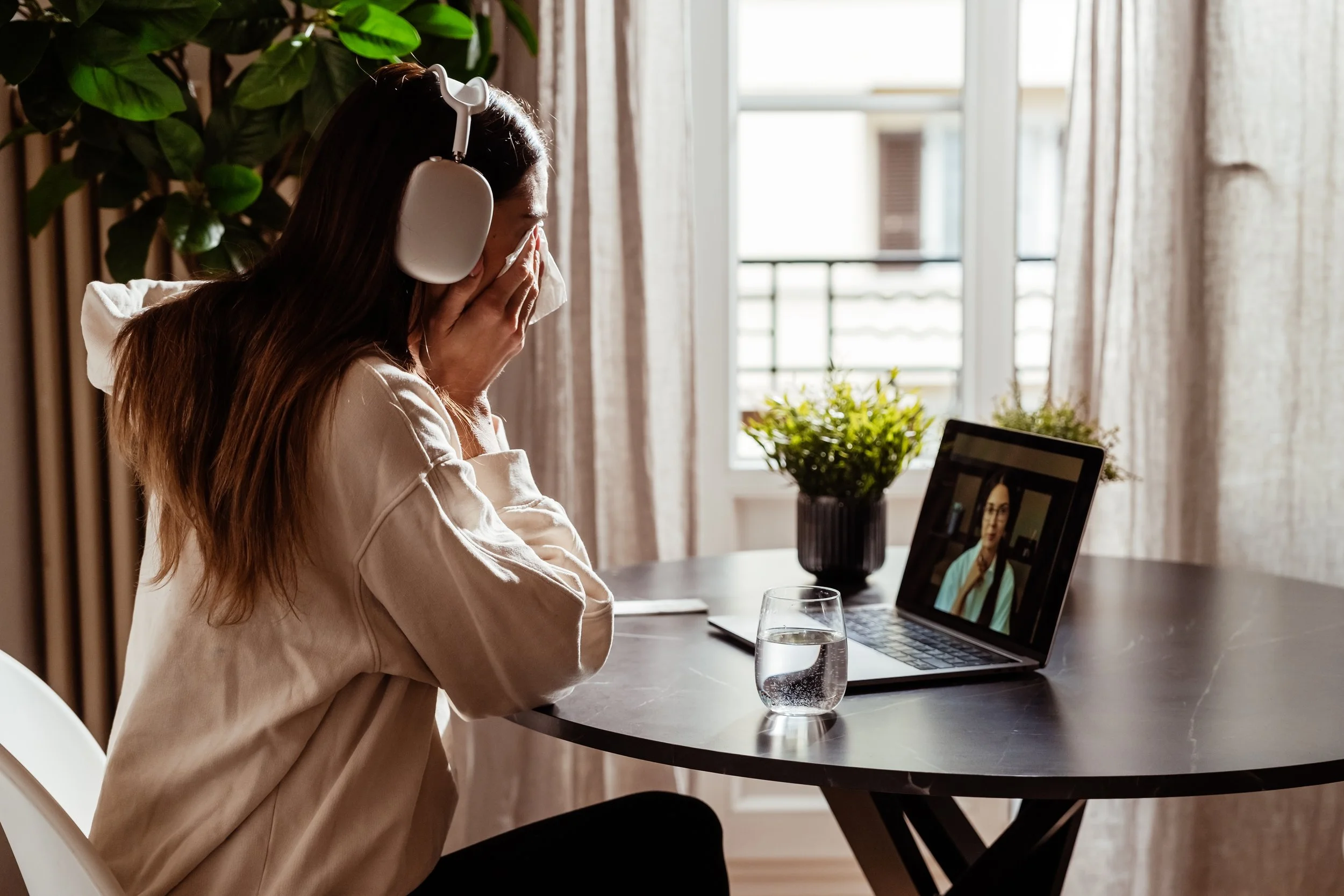 Woman wearing headphones at table, doing online therapy on laptop with a glass of water and potted plant nearby.
