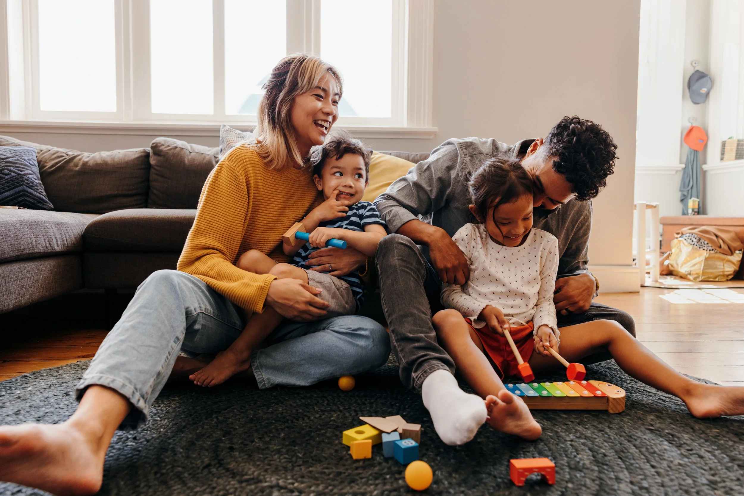 A family of four sitting on the floor of a living room. A woman in a yellow sweater holds a young boy in her lap, while a man in a gray shirt is with a young girl playing a xylophone. The room is bright with a couch and toys scattered around.