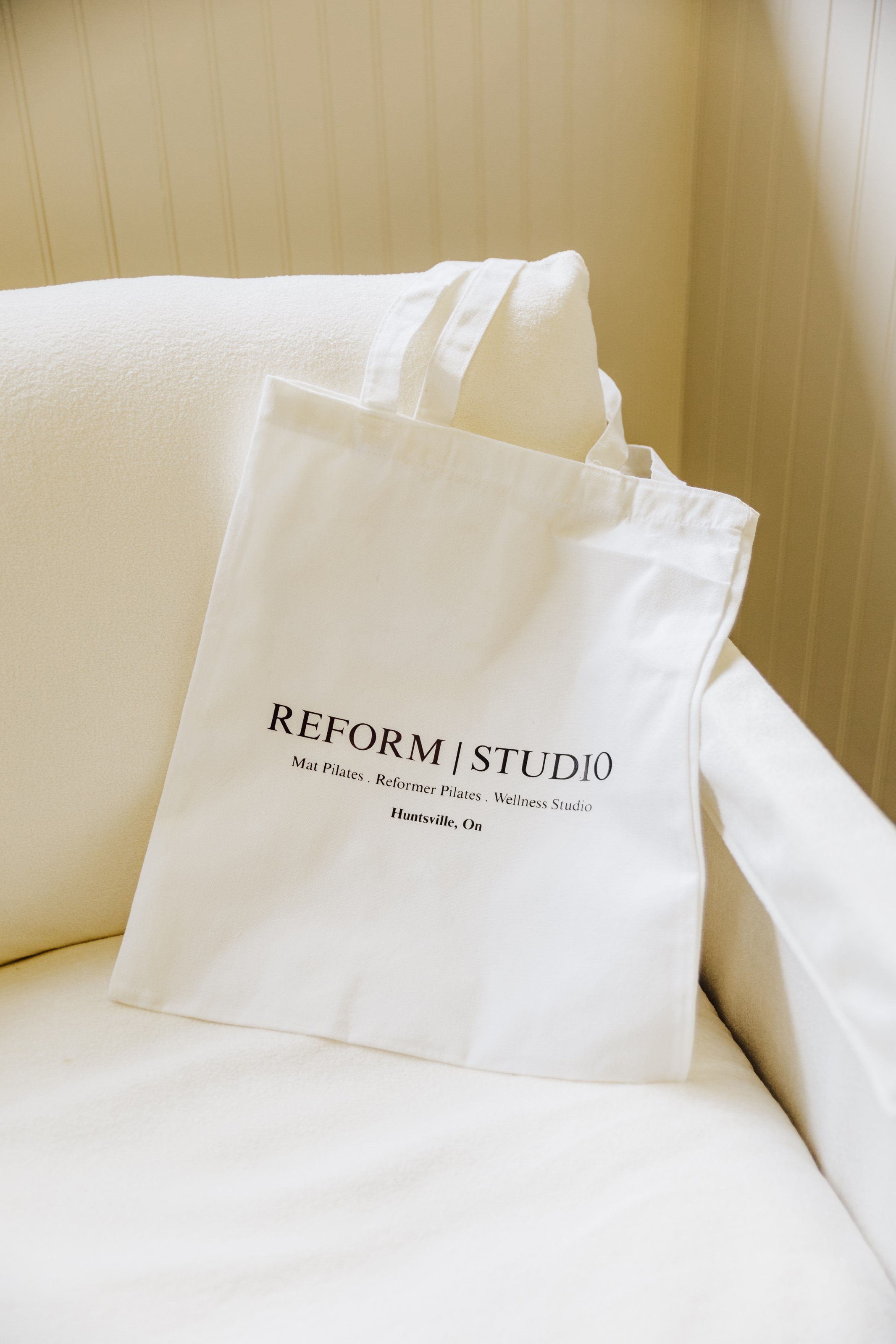 White tote bag with black text reading "REFORM | STUDIO, Mat Pilates, Reformer Pilates, Wellness Studio, Huntsville, ON" resting on a white sofa with a white pillow and beige wall in the background.