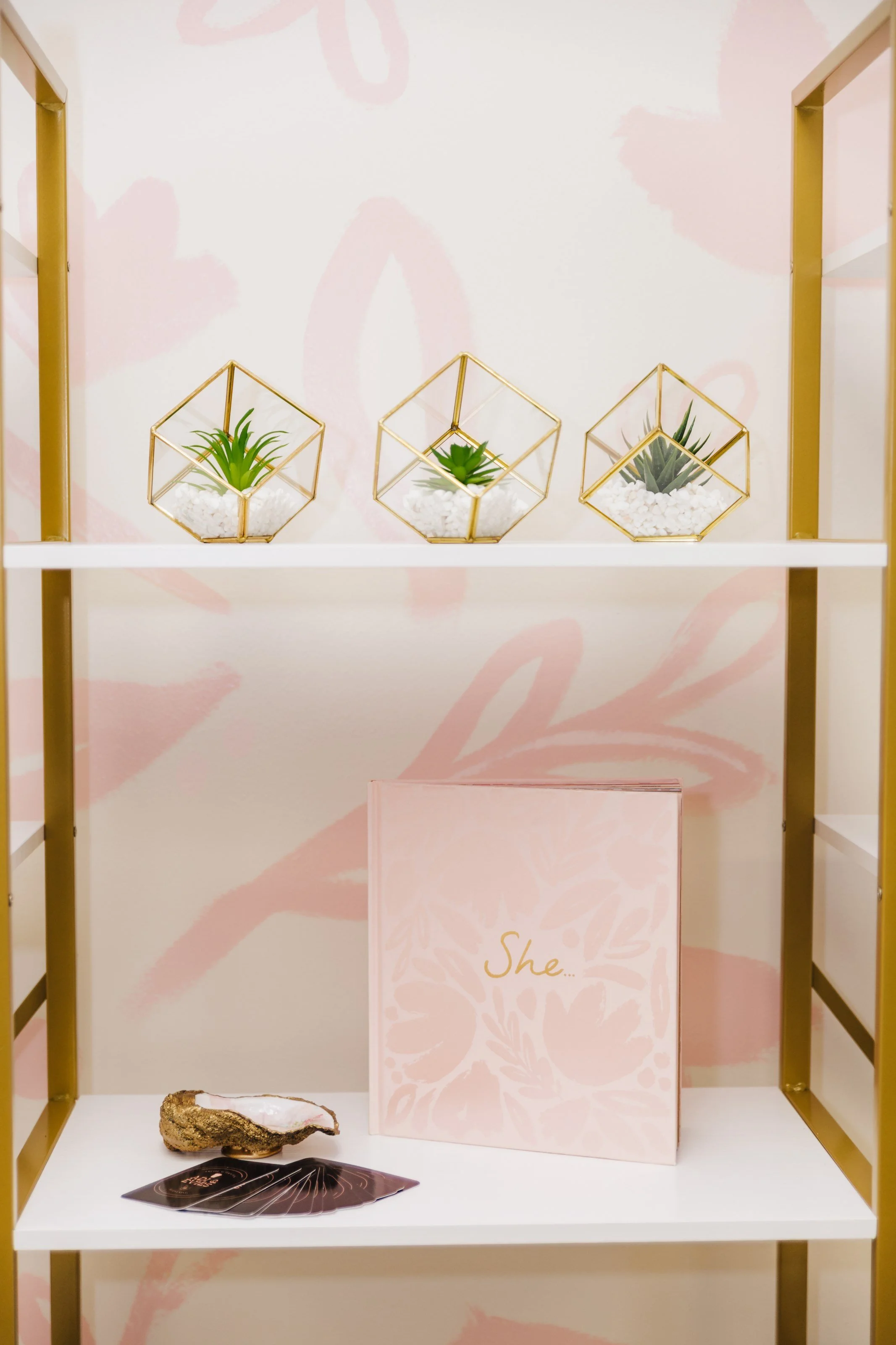Decorative display with three geometric glass planters containing succulents, a pink book with the word 'She' on the cover, a cluster of black cards or coasters, and a piece of geode on white shelves with a light pink and beige floral and brushstroke background.
