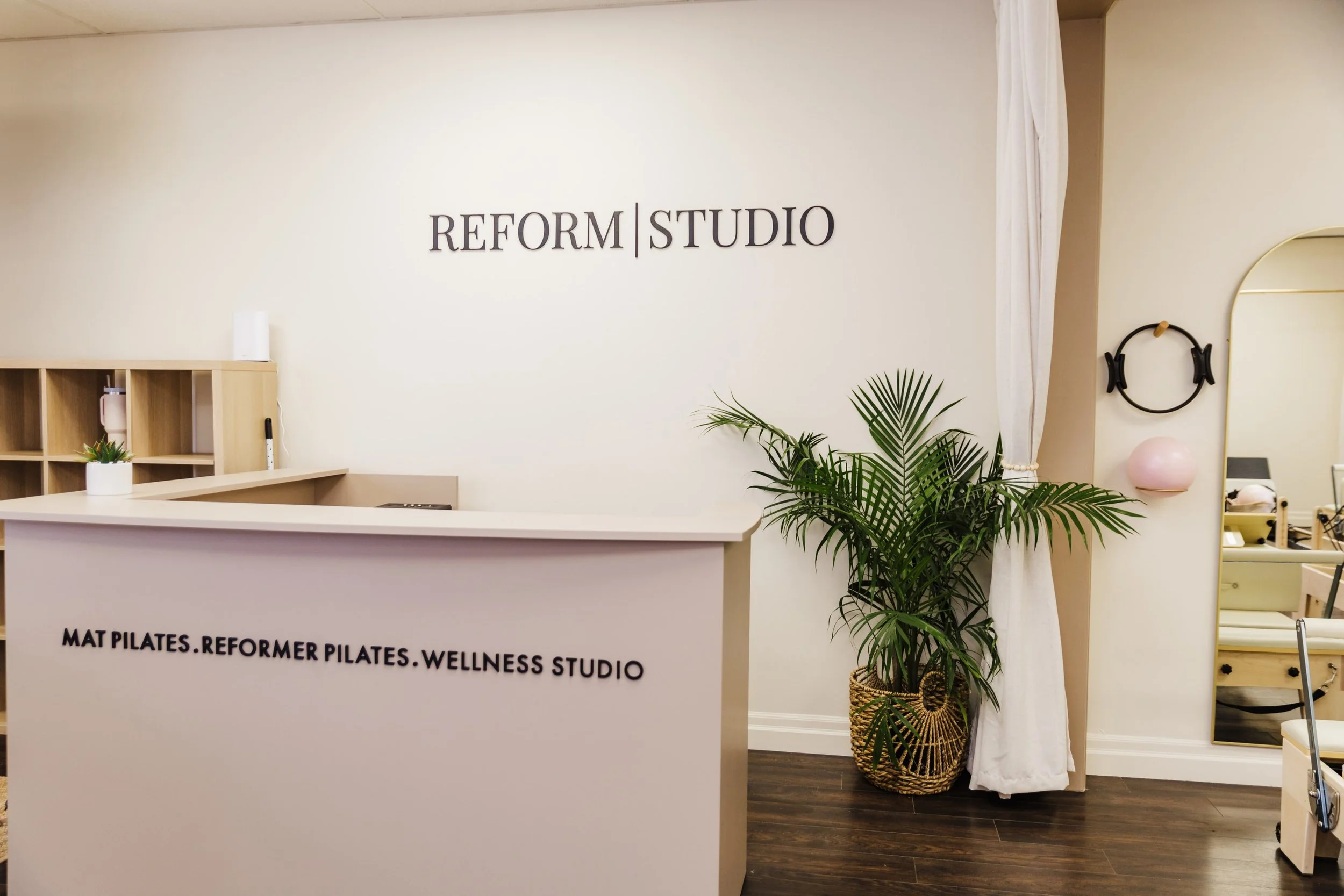 Reception desk with sign reading 'Mat Pilates, Reformer Pilates, Wellness Studio' in a wellness studio, with a potted plant, mirror, and headphones on the wall.