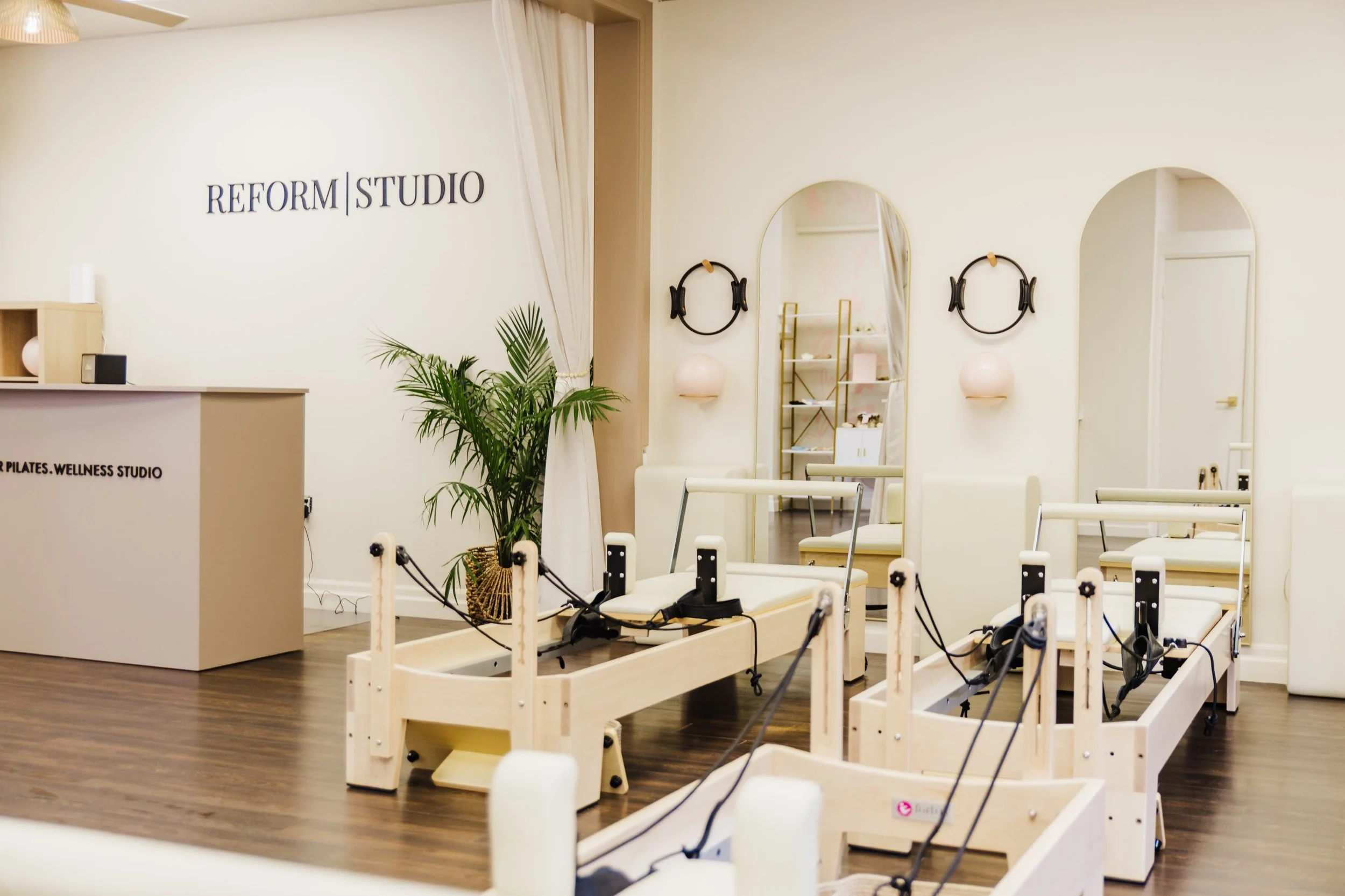 A wellness studio with Pilates reformer machines, a large mirror, and a potted plant in a bright, airy room.