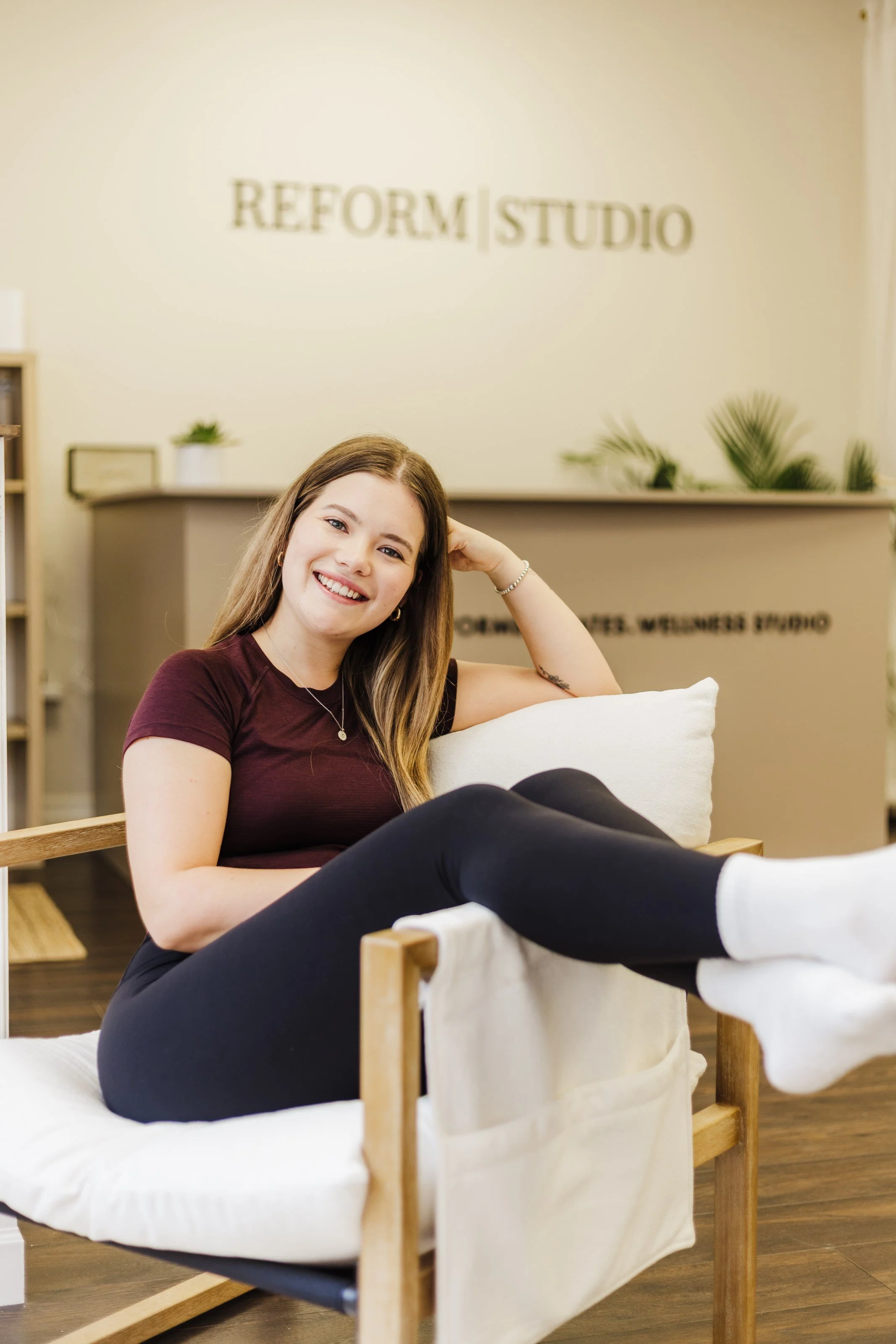 A young woman sitting comfortably on a couch in a wellness studio, smiling at the camera. The studio has a sign in the background that reads 'Reform Studio'.