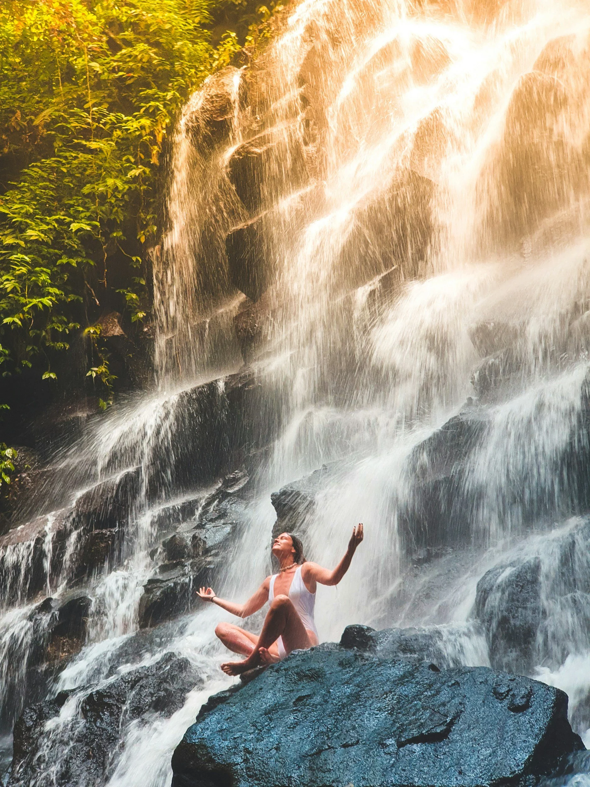 A woman in a white swimsuit sitting cross-legged on a rock under a waterfall with water cascading down from the rocks above, surrounded by green foliage.