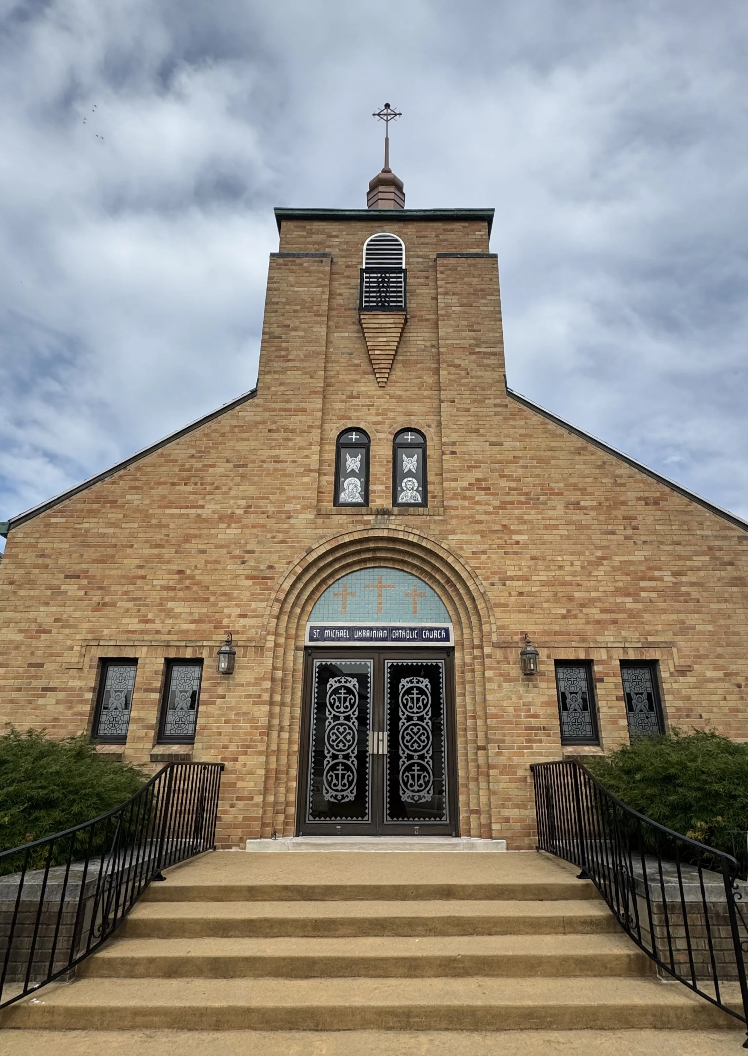 Front view of St. Michael Ukrainian Catholic Church, made of tan brick with black metal doors and steps, arched windows with religious symbols, on a cloudy day with greenery on both sides.